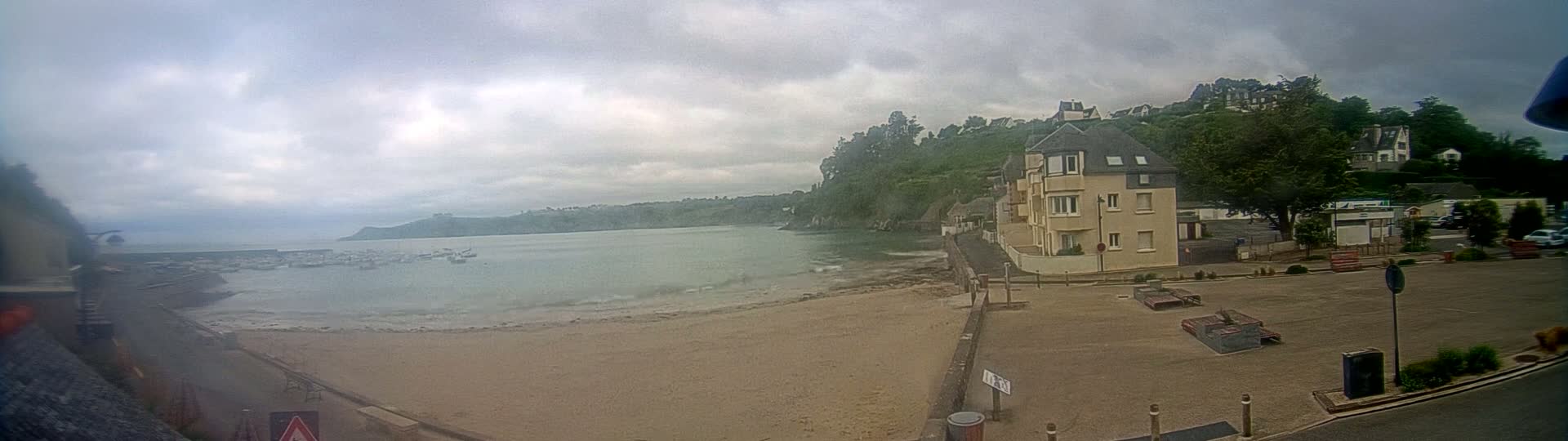 A sandy beach with calm water and several boats is seen next to a coastal town under an overcast sky.