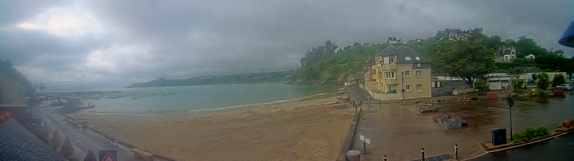A sandy beach and calm bay are seen under an overcast sky, with a multi-story building and other houses situated on a hillside overlooking the water.
