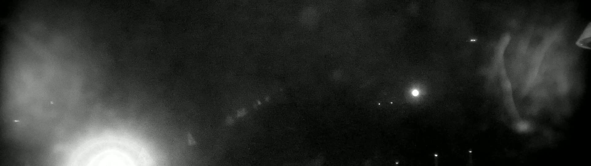 A grainy, low-light image shows a dark, possibly nighttime scene with a few faint lights and hazy, blurry forms in the distance.