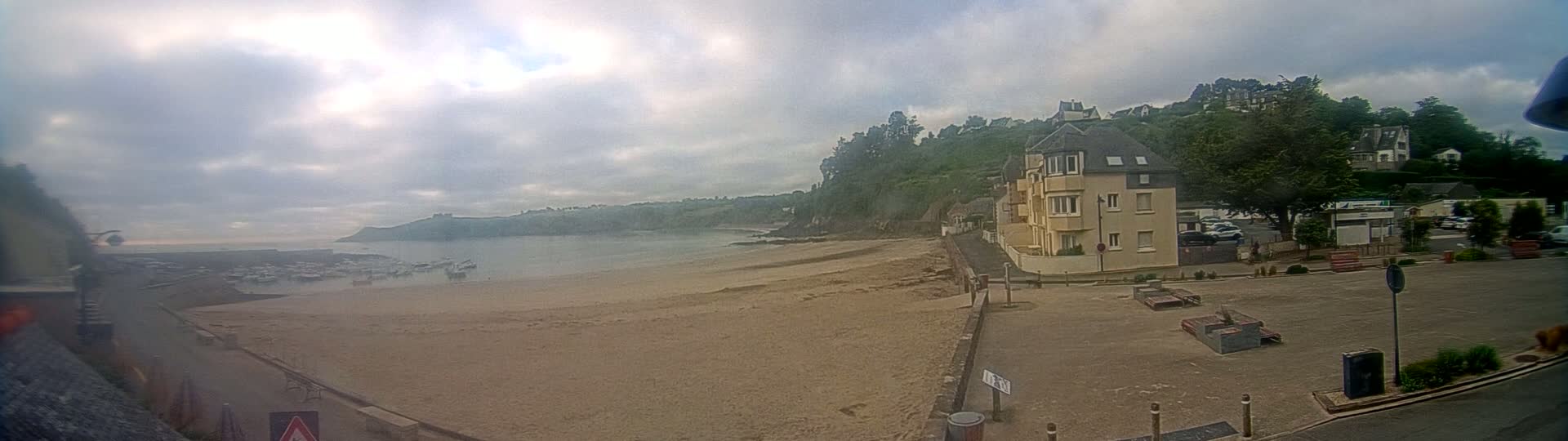A sandy beach stretches below a seaside town under a cloudy sky, with several boats visible in the calm water.