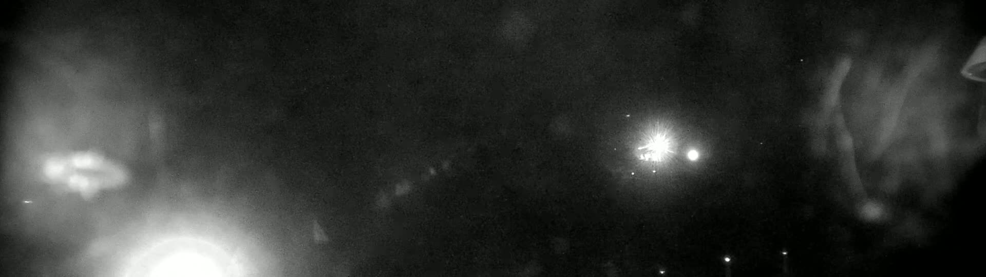 A grainy, black and white night image shows bright lights in the distance and blurry shapes in the foreground, under a dark, possibly cloudy sky.