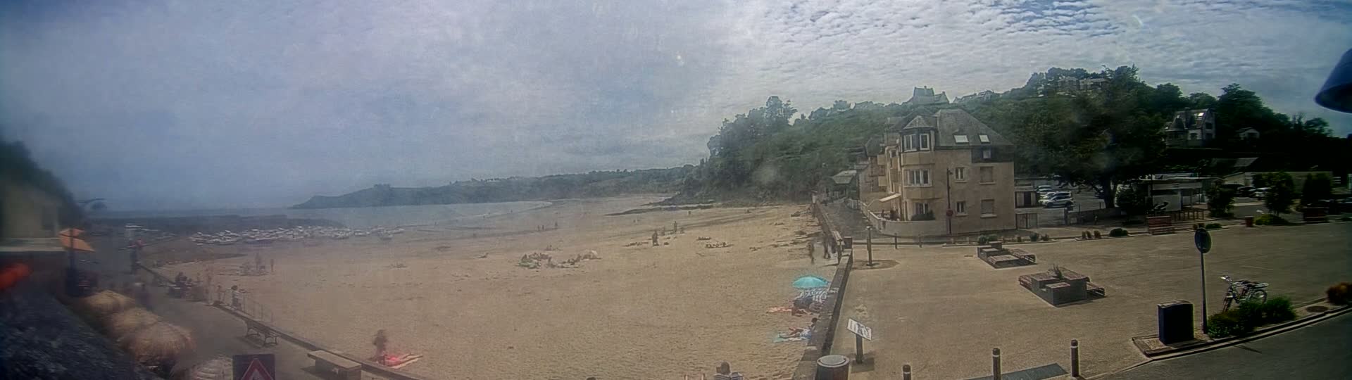 A sandy beach with a few people sunbathing and a building overlooking it on a partly cloudy day.