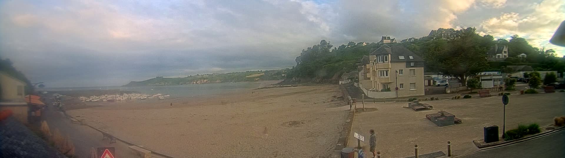 A sandy beach with several boats rests at the edge of calm water, under a cloudy sky, next to a residential area with buildings and parked cars.
