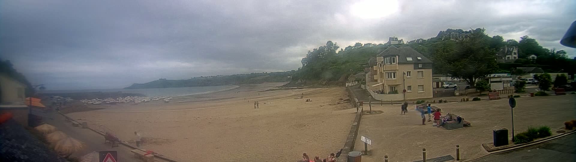 A sandy beach with several small boats in the water is visible under an overcast sky, next to a town with buildings and people.