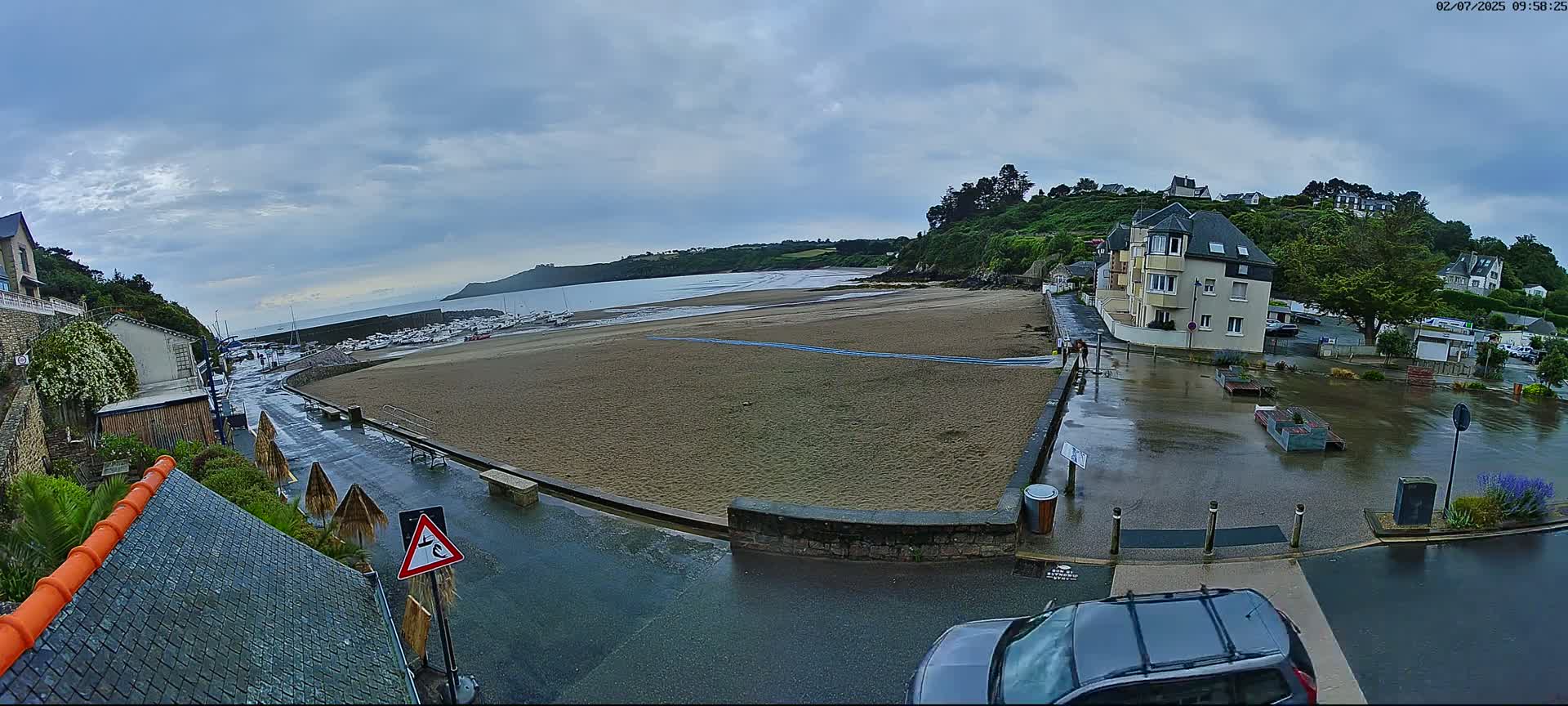 A wet sandy beach, bordered by buildings and a parking lot, is visible under a cloudy sky.