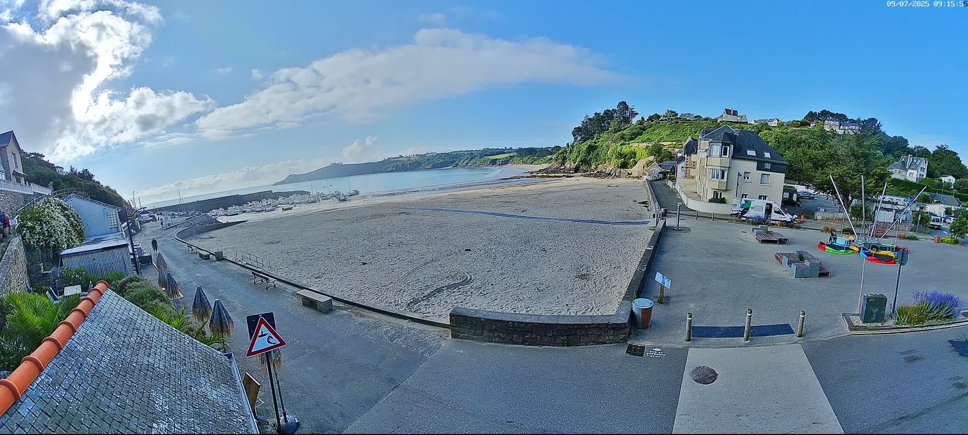 A mostly sunny day reveals a sandy beach with boats in the harbor, buildings on a hillside, and a paved area with some playground equipment.