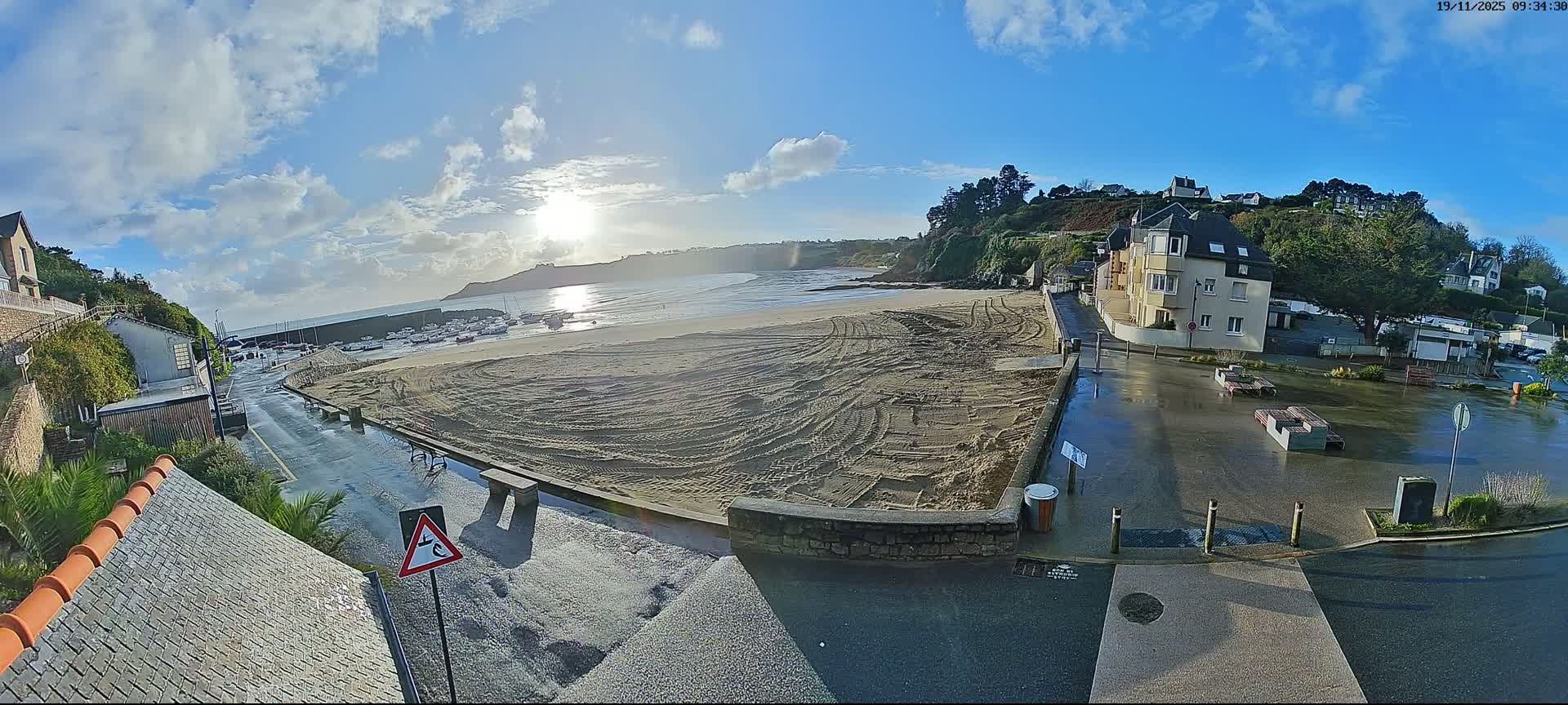 A sunny and partly cloudy morning illuminates a wide coastal scene featuring a sandy beach with tire tracks, a harbor filled with boats, wet streets, and buildings nestled on a tree-covered hillside.