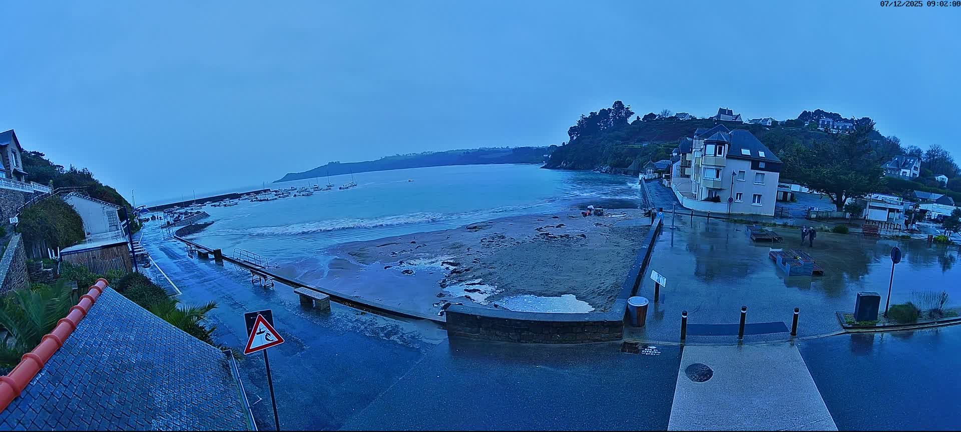 A wide shot captures a coastal town on a damp, overcast day, featuring a wet beach meeting a bay dotted with boats, and buildings nestled along the reflective shoreline and up the surrounding hills.