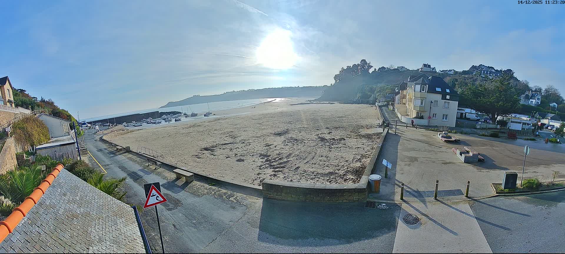 A wide shot captures a coastal town on a damp, overcast day, featuring a wet beach meeting a bay dotted with boats, and buildings nestled along the reflective shoreline and up the surrounding hills.