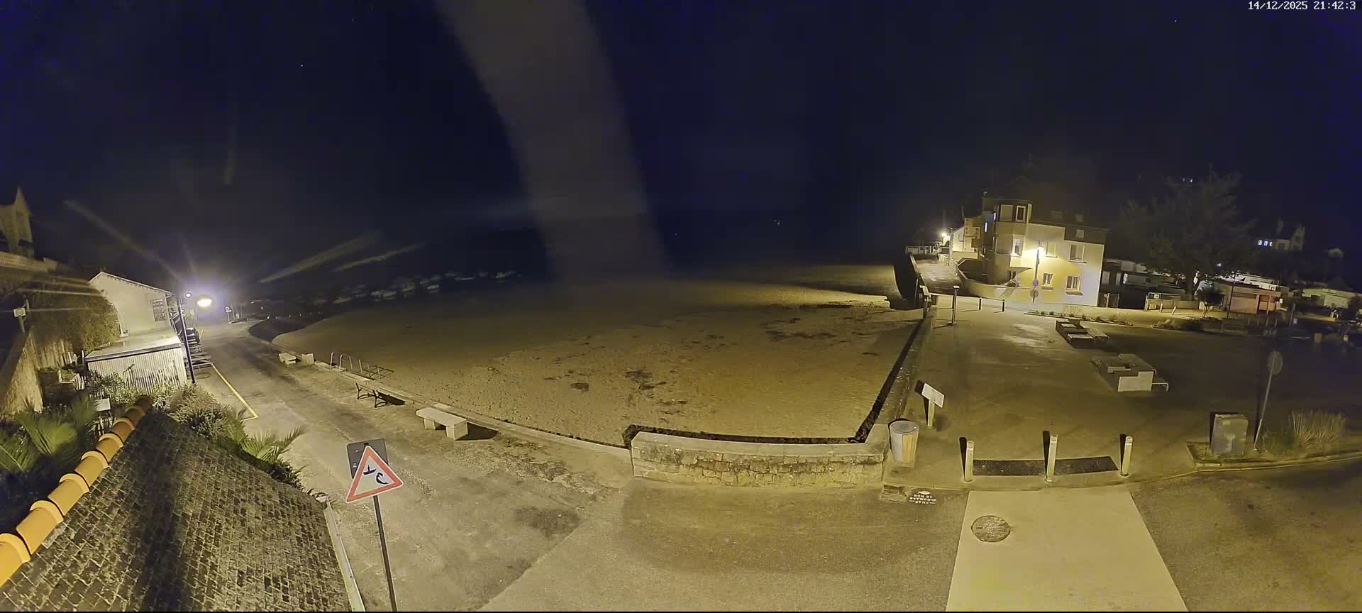 A wide shot captures a coastal town on a damp, overcast day, featuring a wet beach meeting a bay dotted with boats, and buildings nestled along the reflective shoreline and up the surrounding hills.