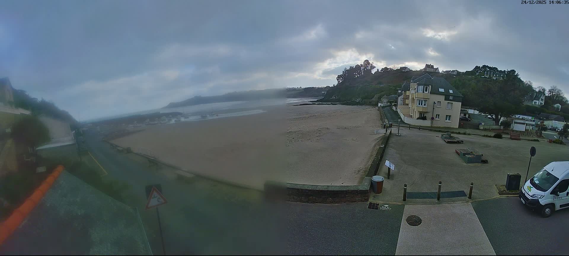 A wide shot captures a coastal town on a damp, overcast day, featuring a wet beach meeting a bay dotted with boats, and buildings nestled along the reflective shoreline and up the surrounding hills.