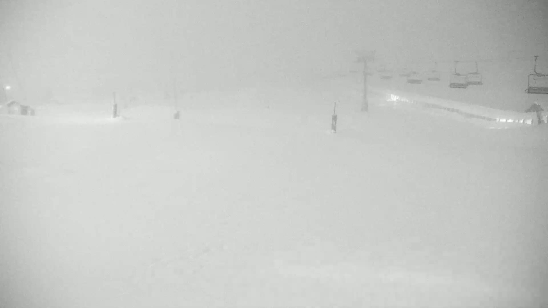 A snow-covered ski slope is mostly obscured by heavy snowfall, with chairlifts faintly visible in the distance.
