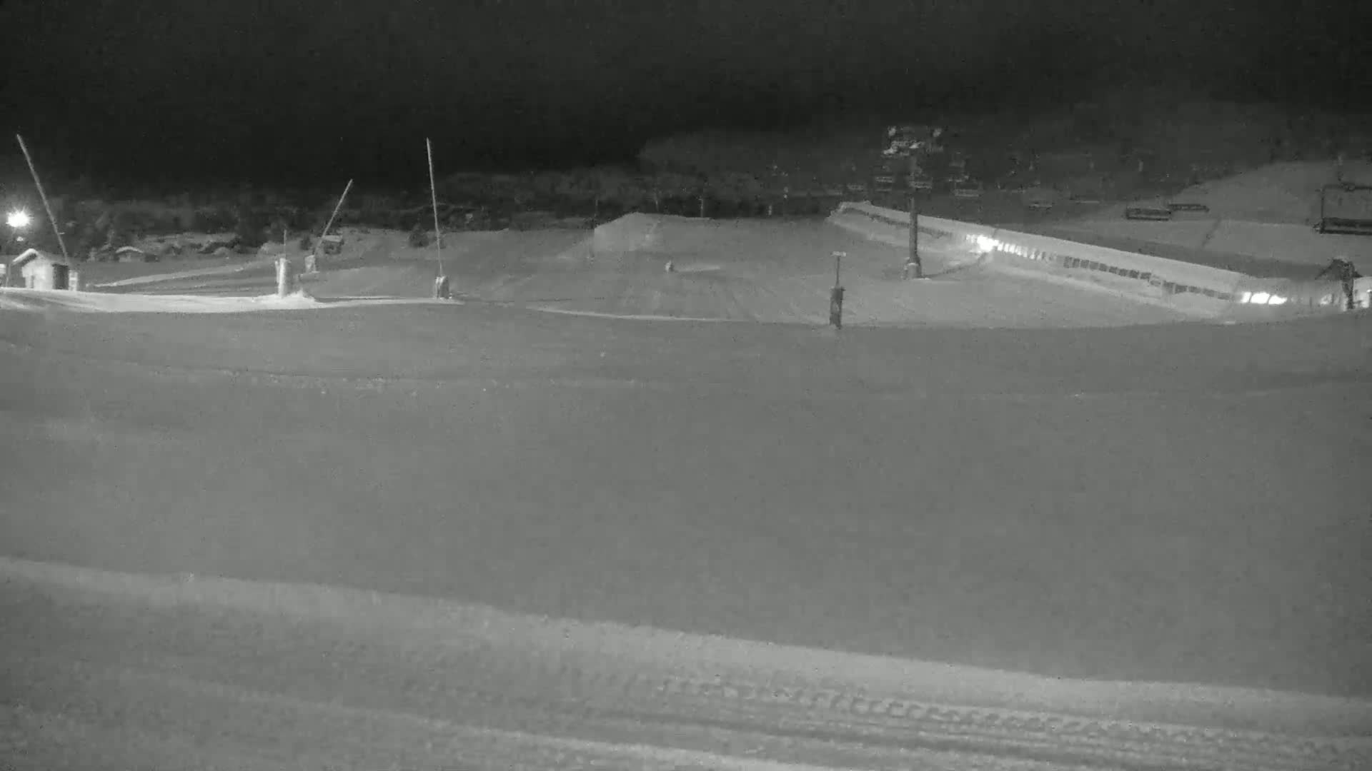 A snow-covered ski slope at night, illuminated by artificial lights, features ski lifts and small buildings.