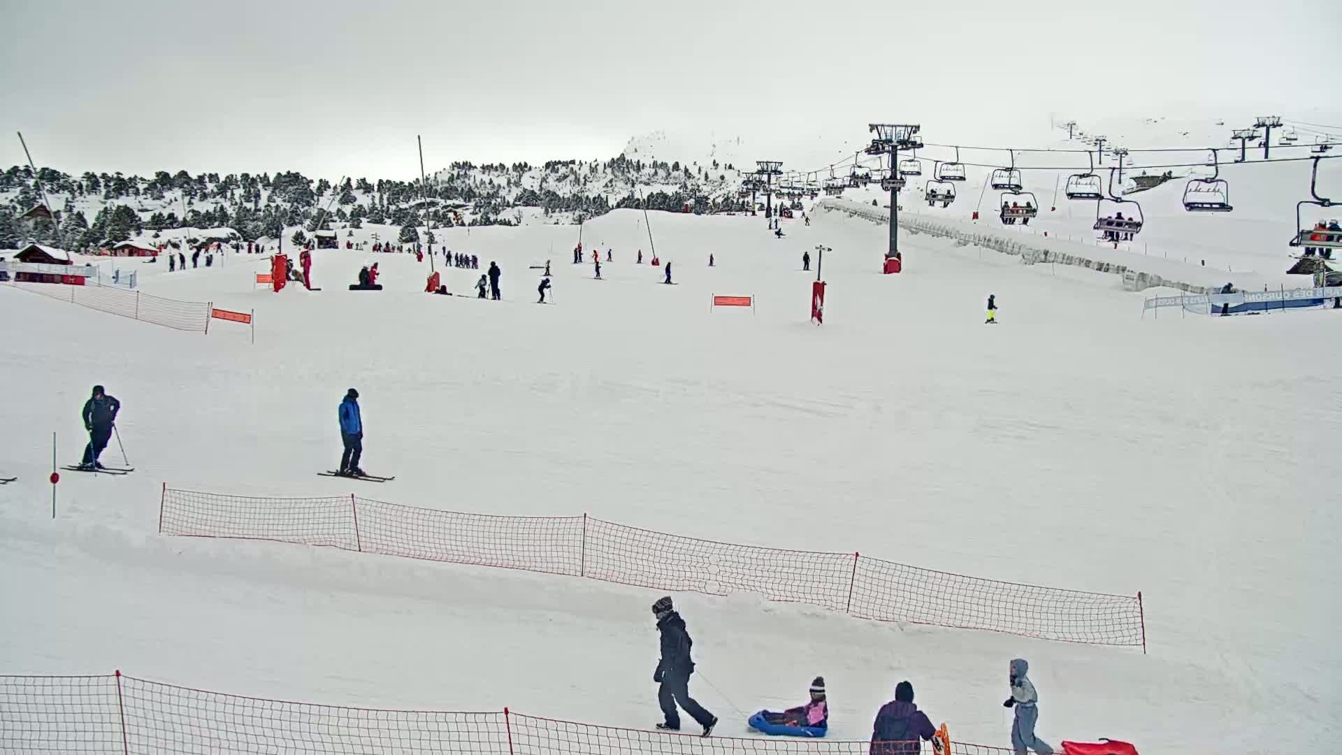 A snowy ski slope with several skiers and snowboarders, a chairlift, and people sledding under an overcast sky.