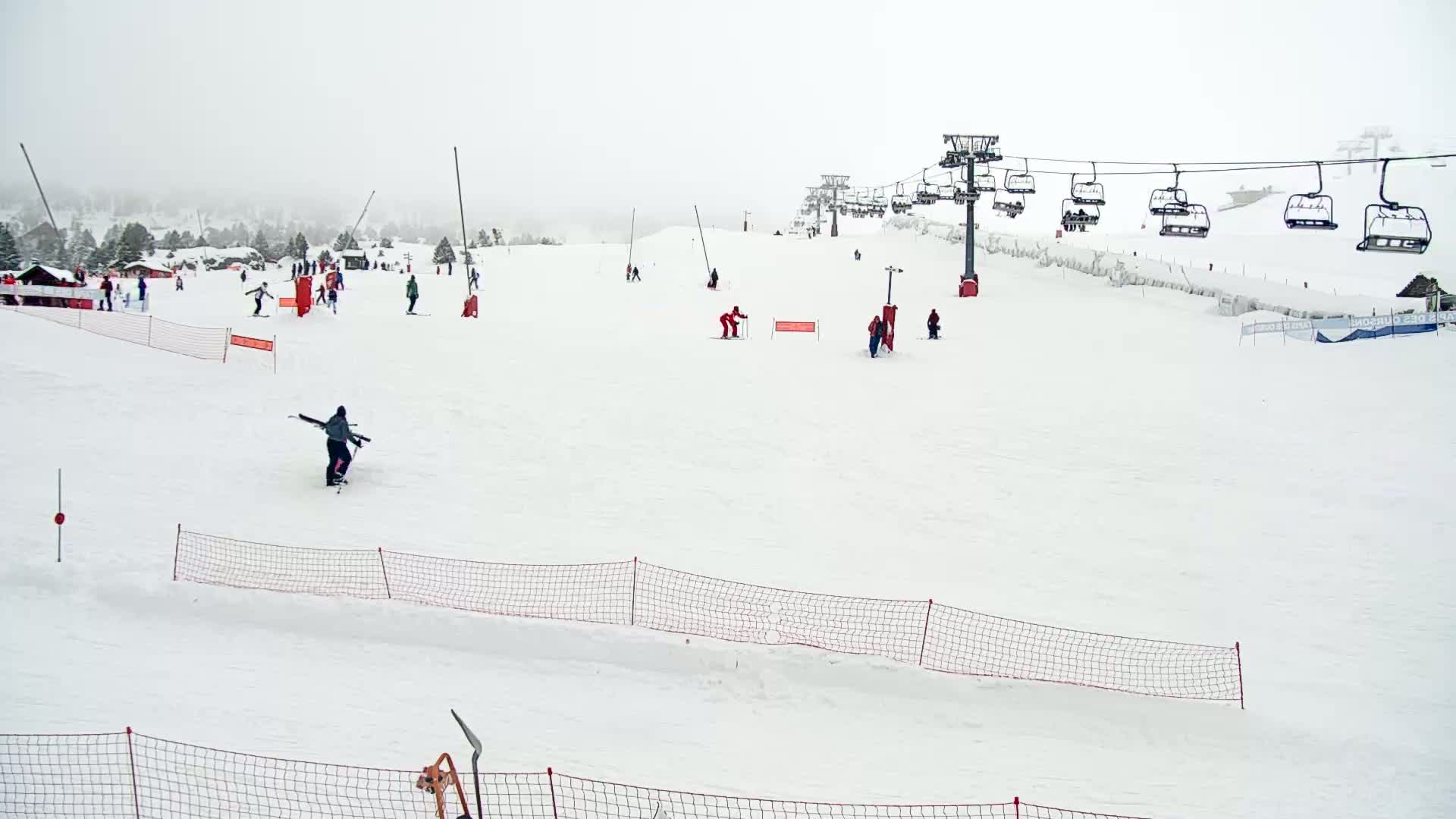 A snowy ski slope with several skiers and snowboarders, chairlifts, and safety nets under overcast skies.