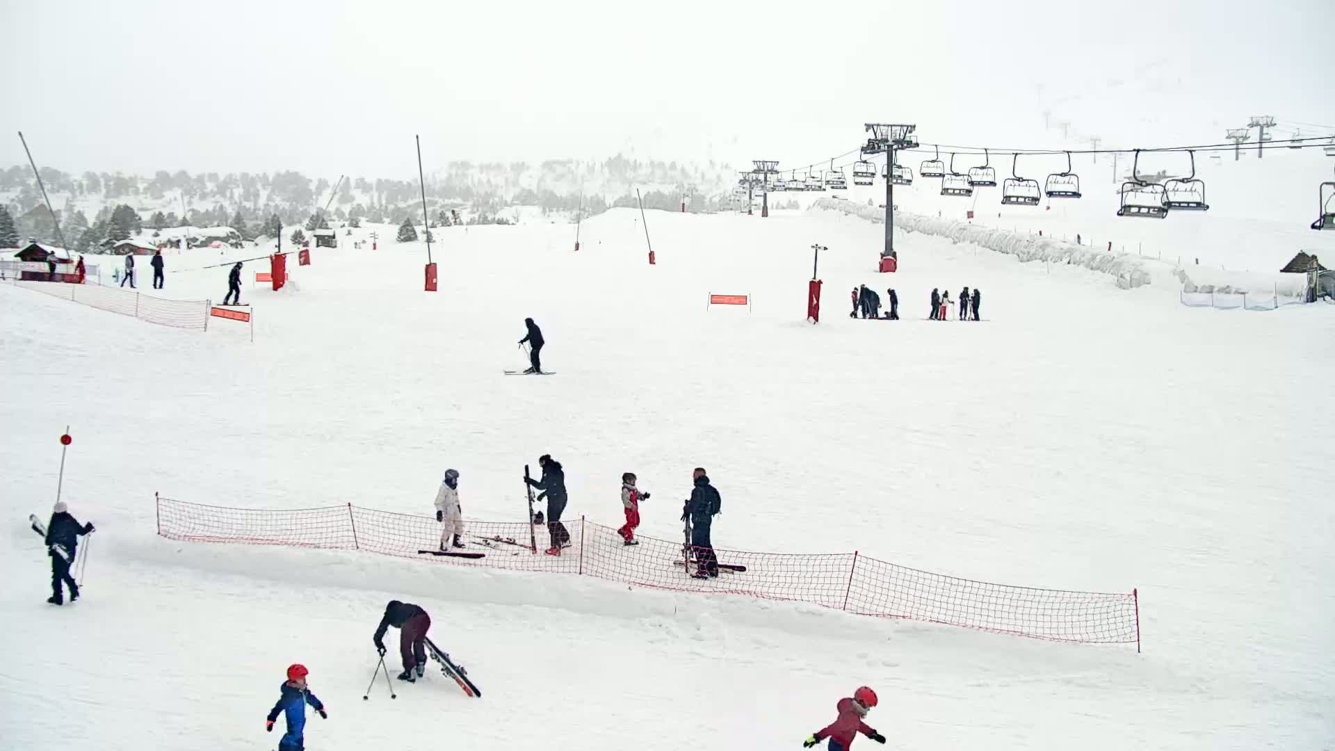 A snowy ski slope with several skiers and snowboarders, a chairlift, and a few small buildings in the background under overcast skies.
