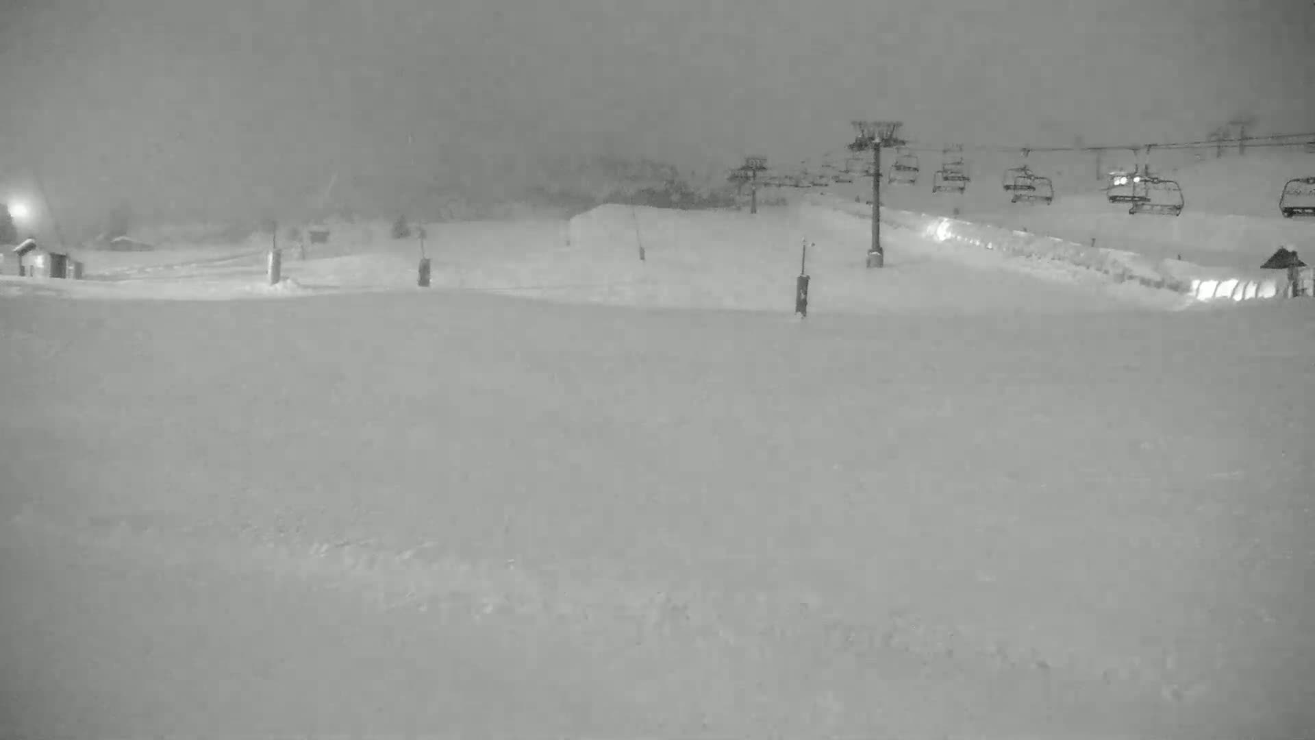 A snow-covered ski resort at night is seen with a chairlift and several chairs visible in the background under heavy snow.