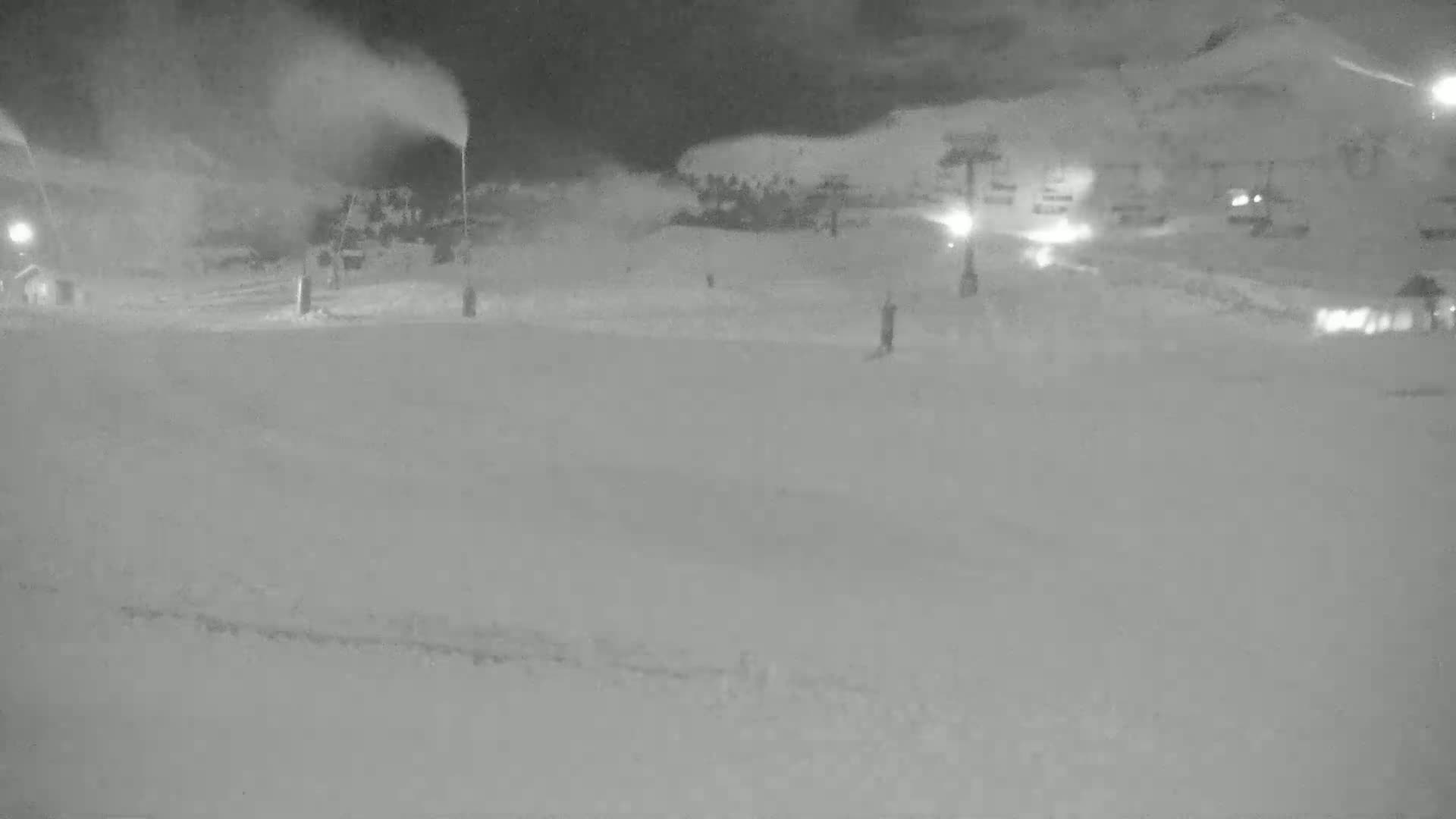 A snowmaking machine sprays snow onto a snowy, nighttime ski slope with buildings visible in the background.