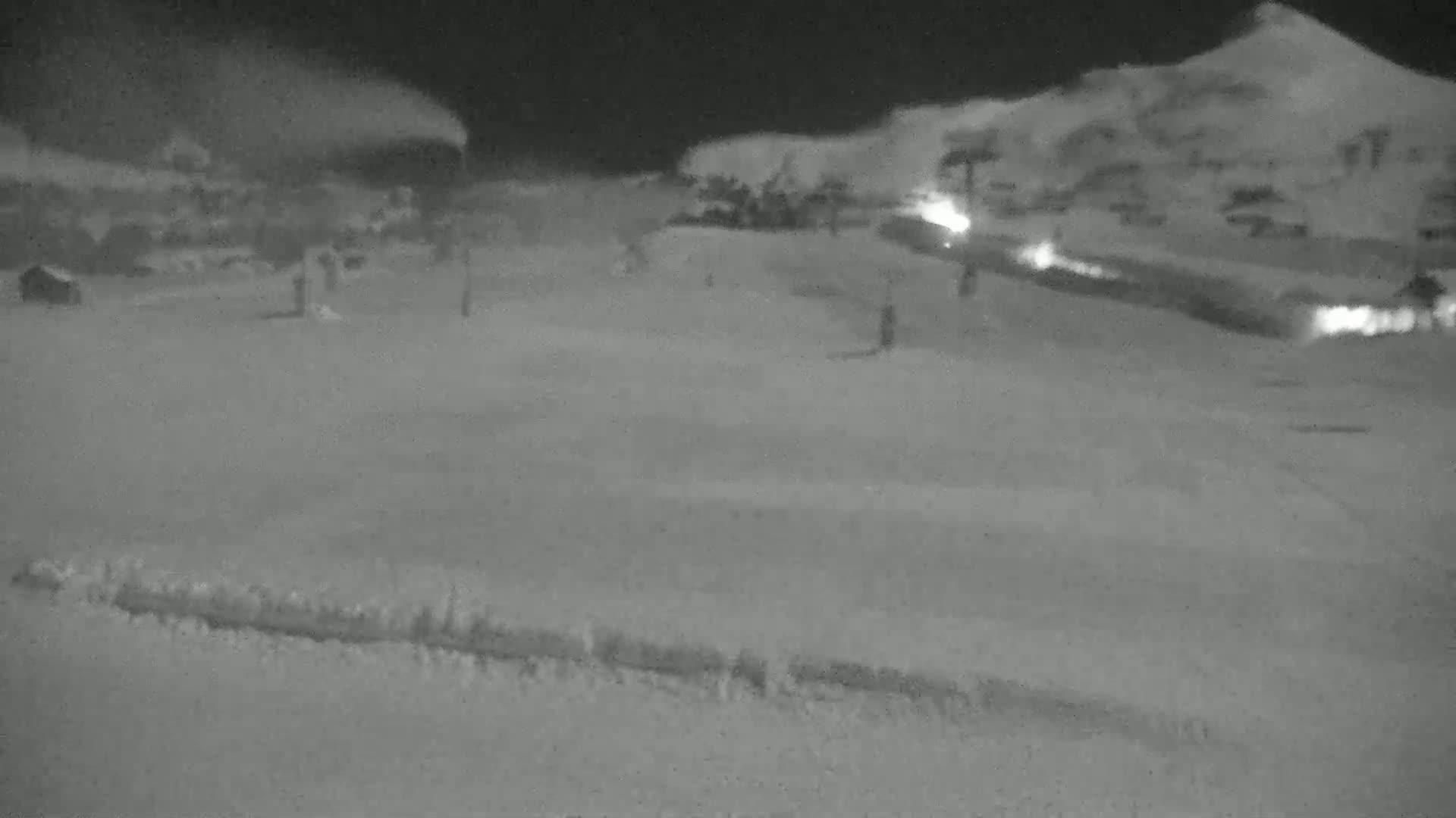 A snow-covered landscape at night, with blowing snow and faint lights visible in the distance.