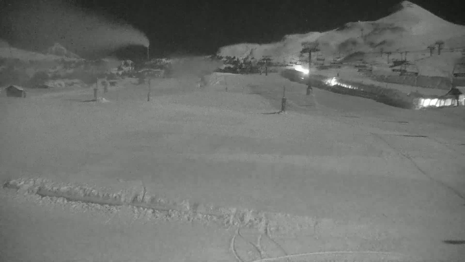 A snow-covered landscape at night, with snow blowing from snowmaking machines near ski lifts and buildings in the background.