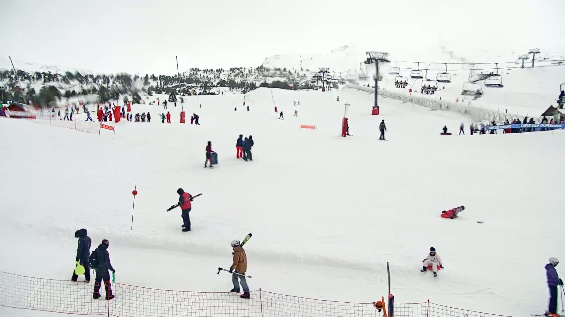 A snowy ski slope with several people skiing, snowboarding, and sledding, under an overcast sky.