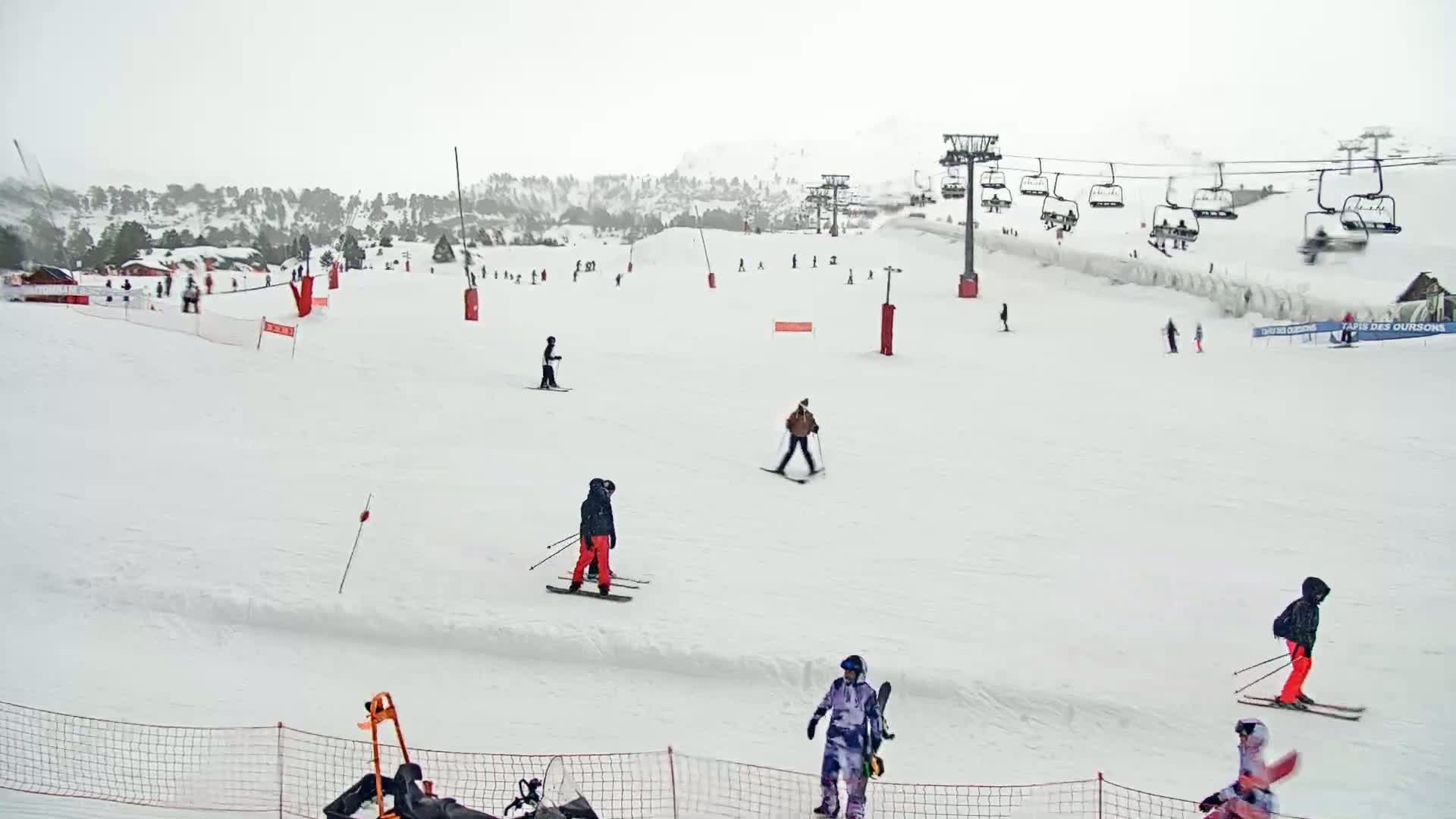 A snowy ski slope with several skiers and snowboarders, chairlifts, and some small buildings in the background under an overcast sky.