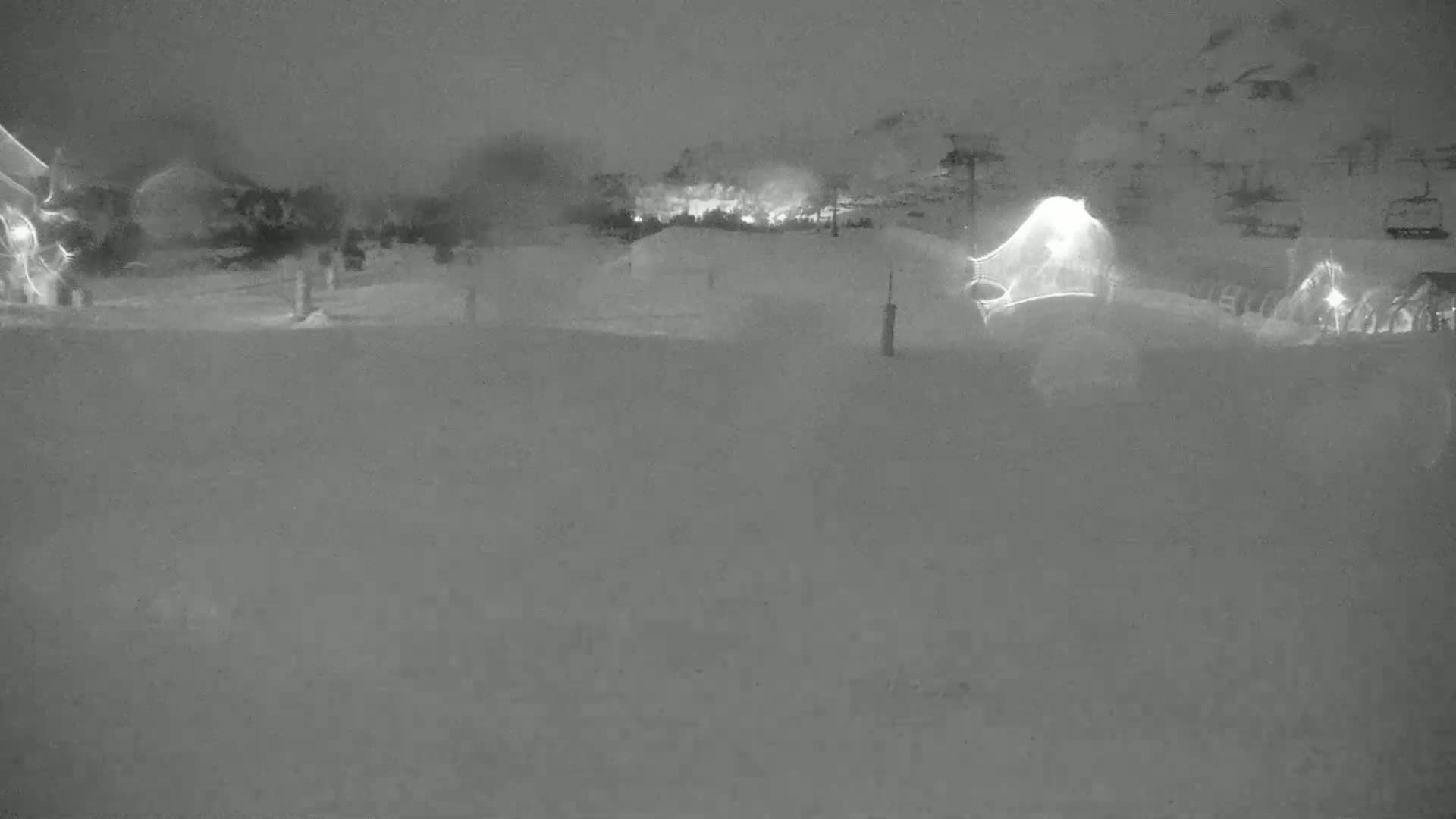 A snow-covered landscape at night, illuminated by faint lights in the distance, with a ski lift visible.