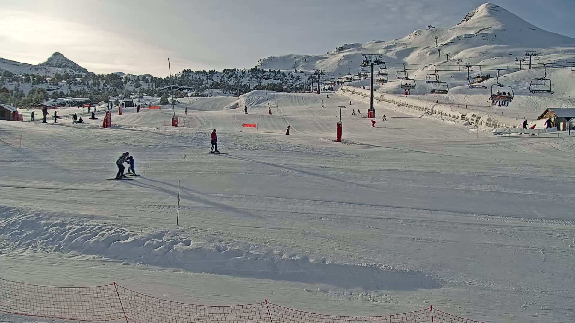A mostly sunny ski slope with several skiers and snowboarders, chairlifts, and snow-covered mountains in the background.