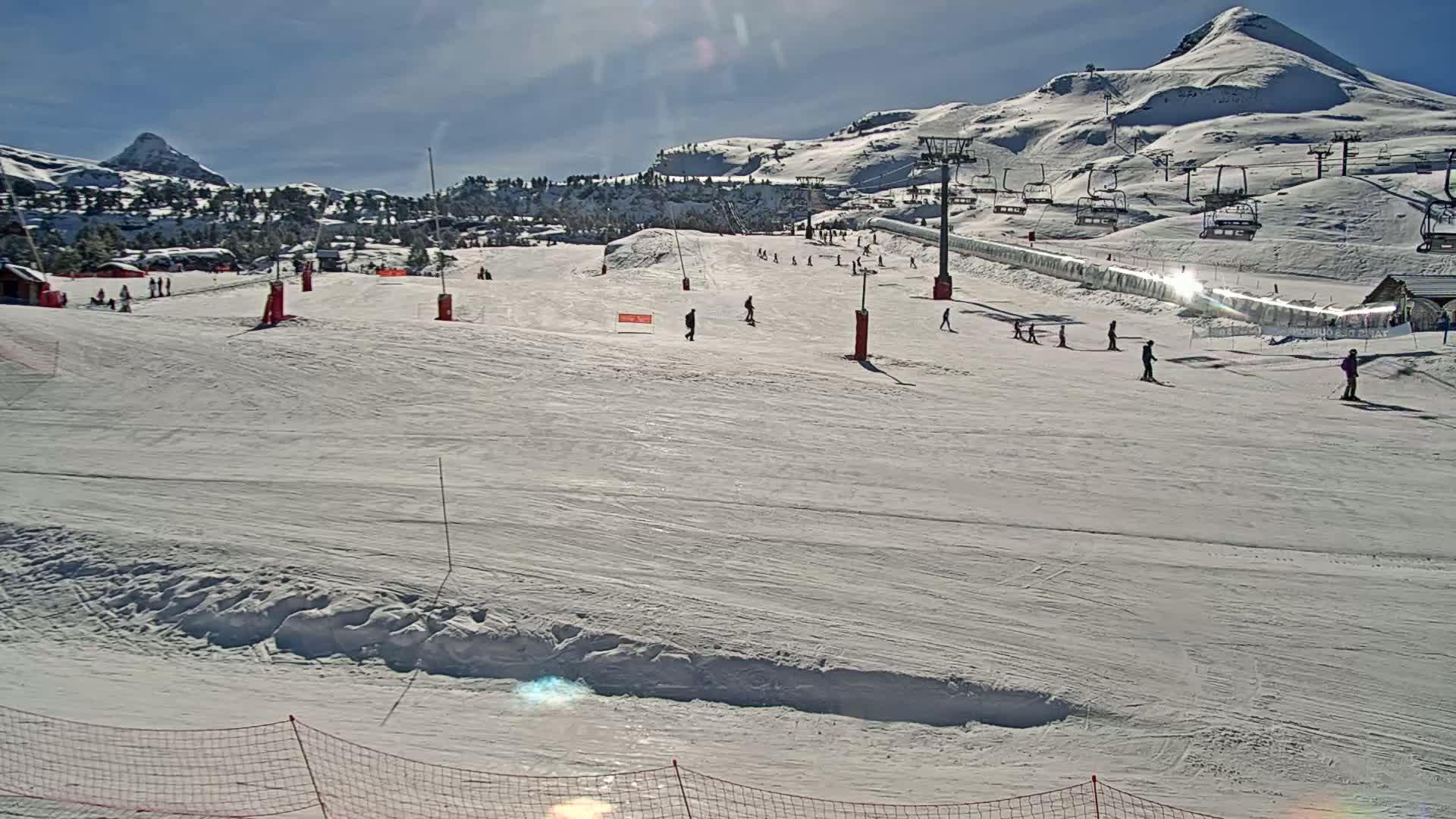 A snowy ski slope with several skiers and snowboarders, ski lifts, and snow-covered mountains in the background under sunny skies.
