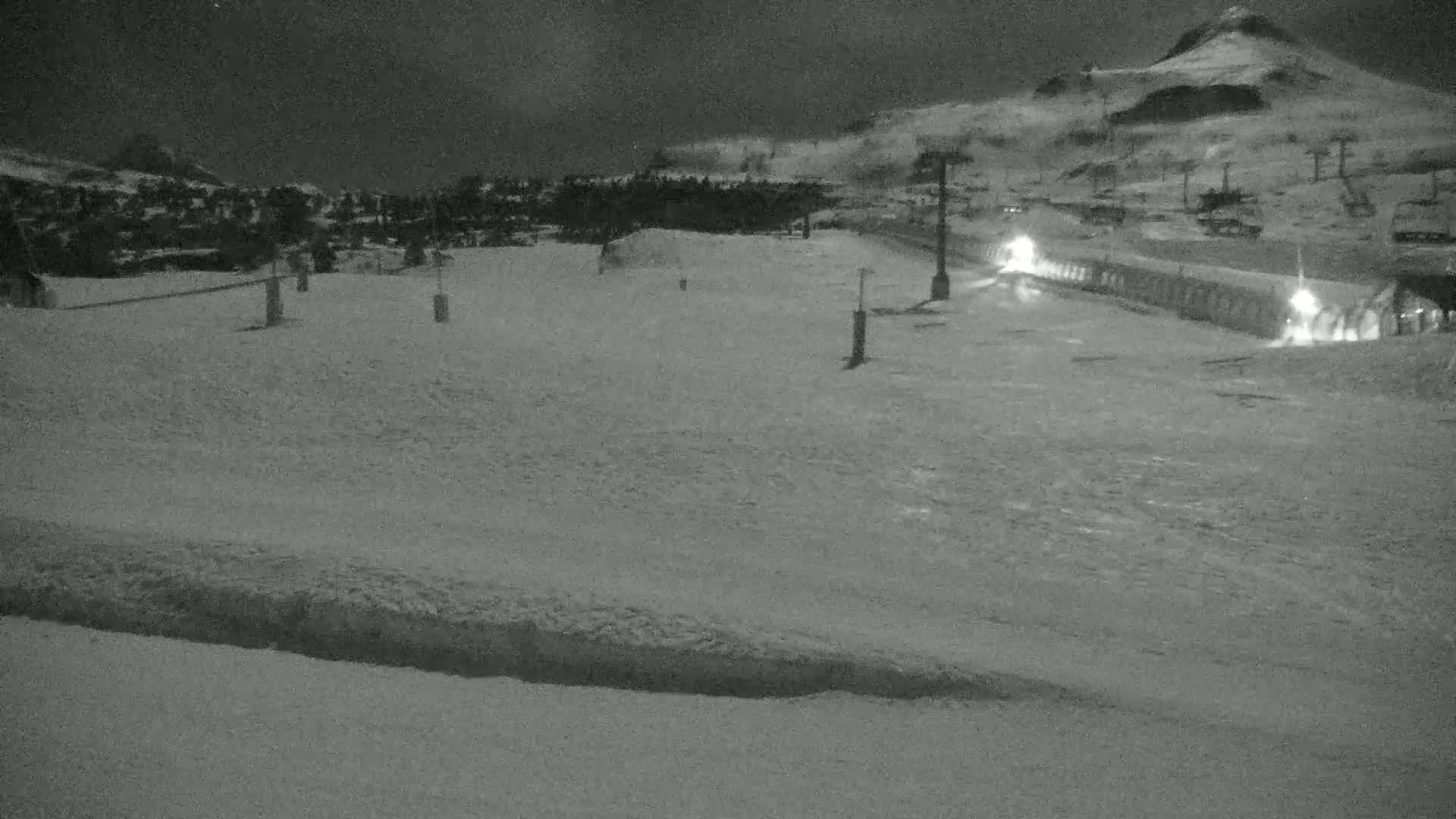A snow-covered ski resort at night, with ski lifts and a building illuminated, under a dark sky.