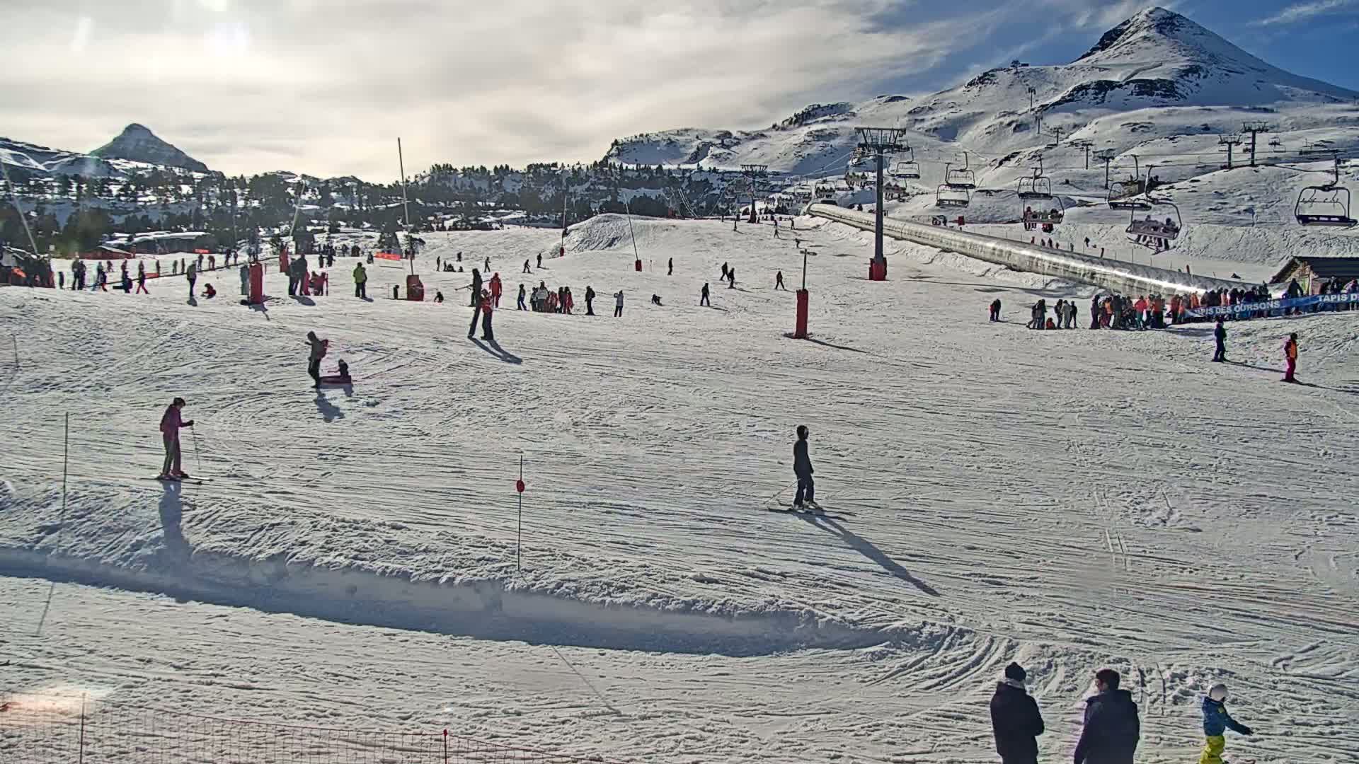 A sunny day on a snow-covered ski slope shows numerous skiers and snowboarders enjoying the mountainside, with ski lifts and a mountain peak in the background.