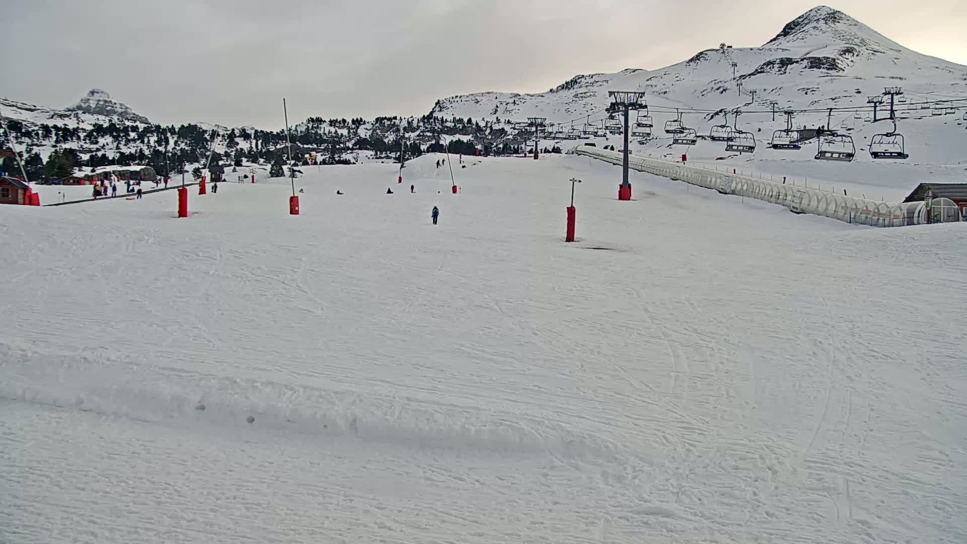 A snowy ski slope with a chairlift and a few skiers and snowboarders under an overcast sky.