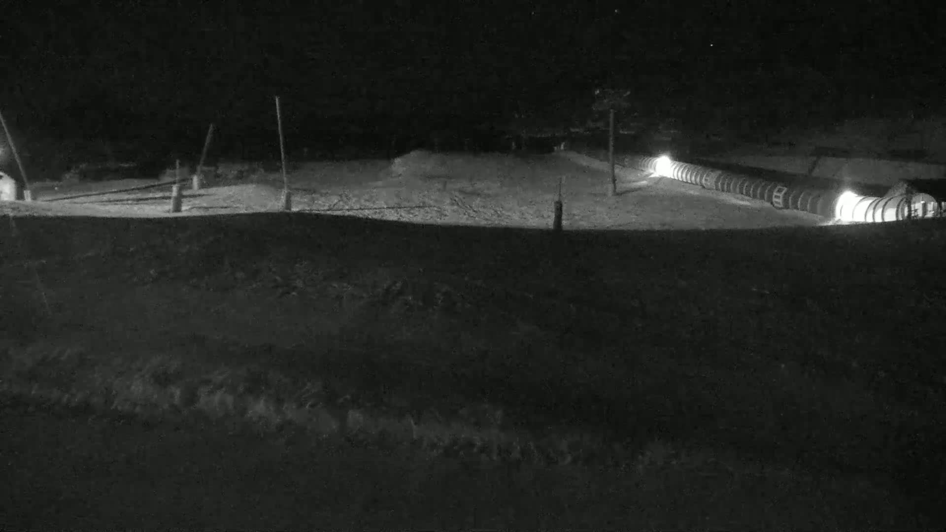 A snow-covered, dimly lit area at night shows a long, illuminated structure resembling a series of connected pipes or cylinders.