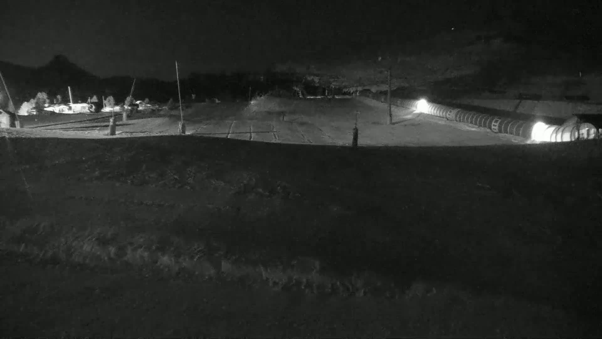 A snow-covered area at night, illuminated by lights at the bottom right, with a long, dark structure, possibly a snowmaking device.