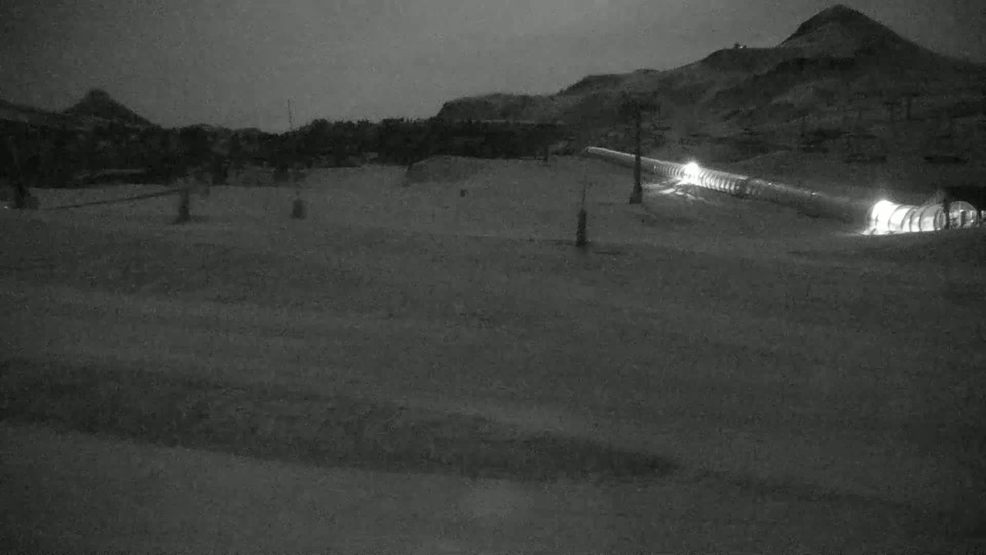 A snow-covered landscape at night is illuminated by the lights of a ski lift extending towards a hill in the background.