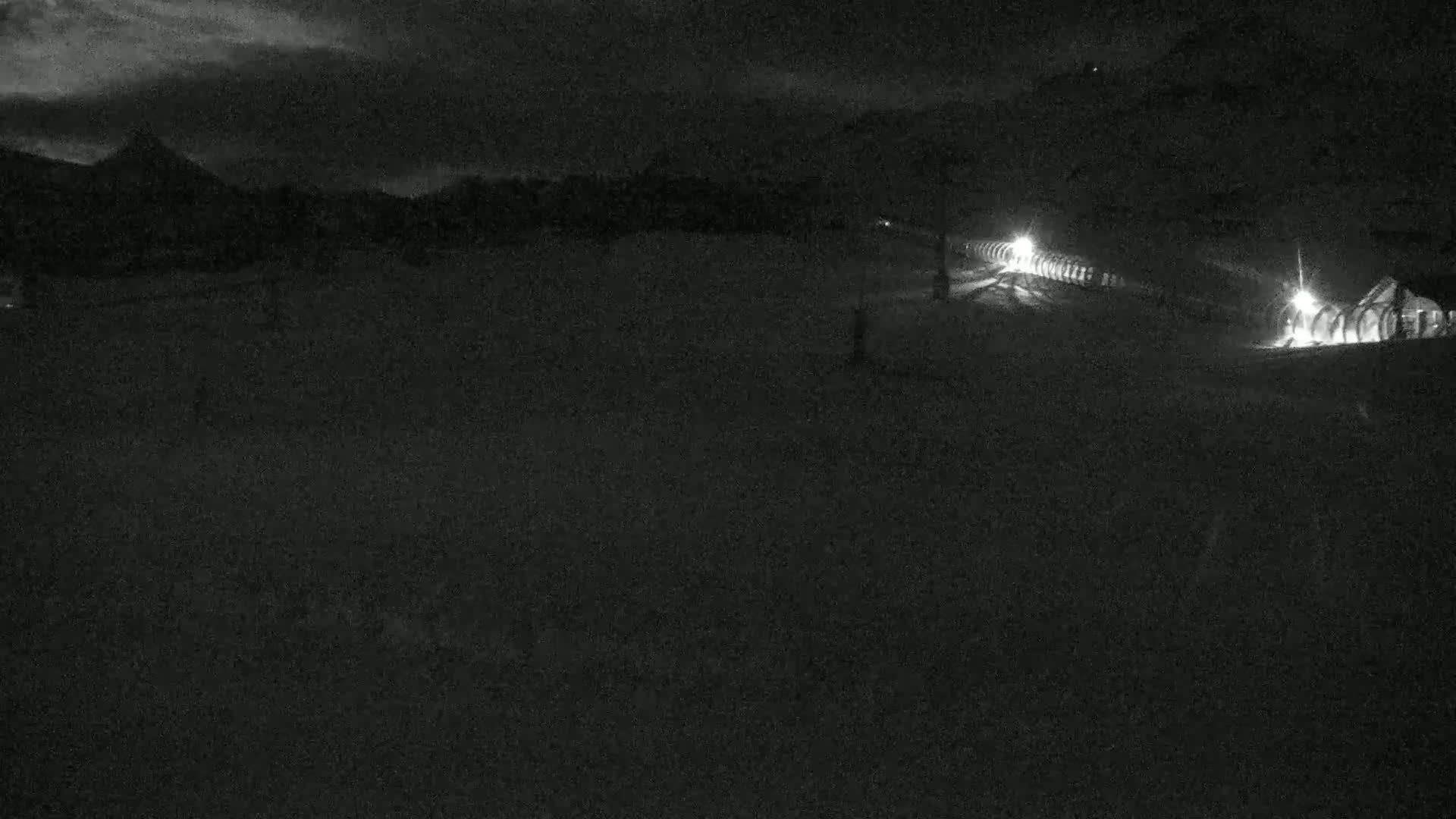 A nighttime, snowy landscape is illuminated by several lights near a building in the distance under a dark, cloudy sky.