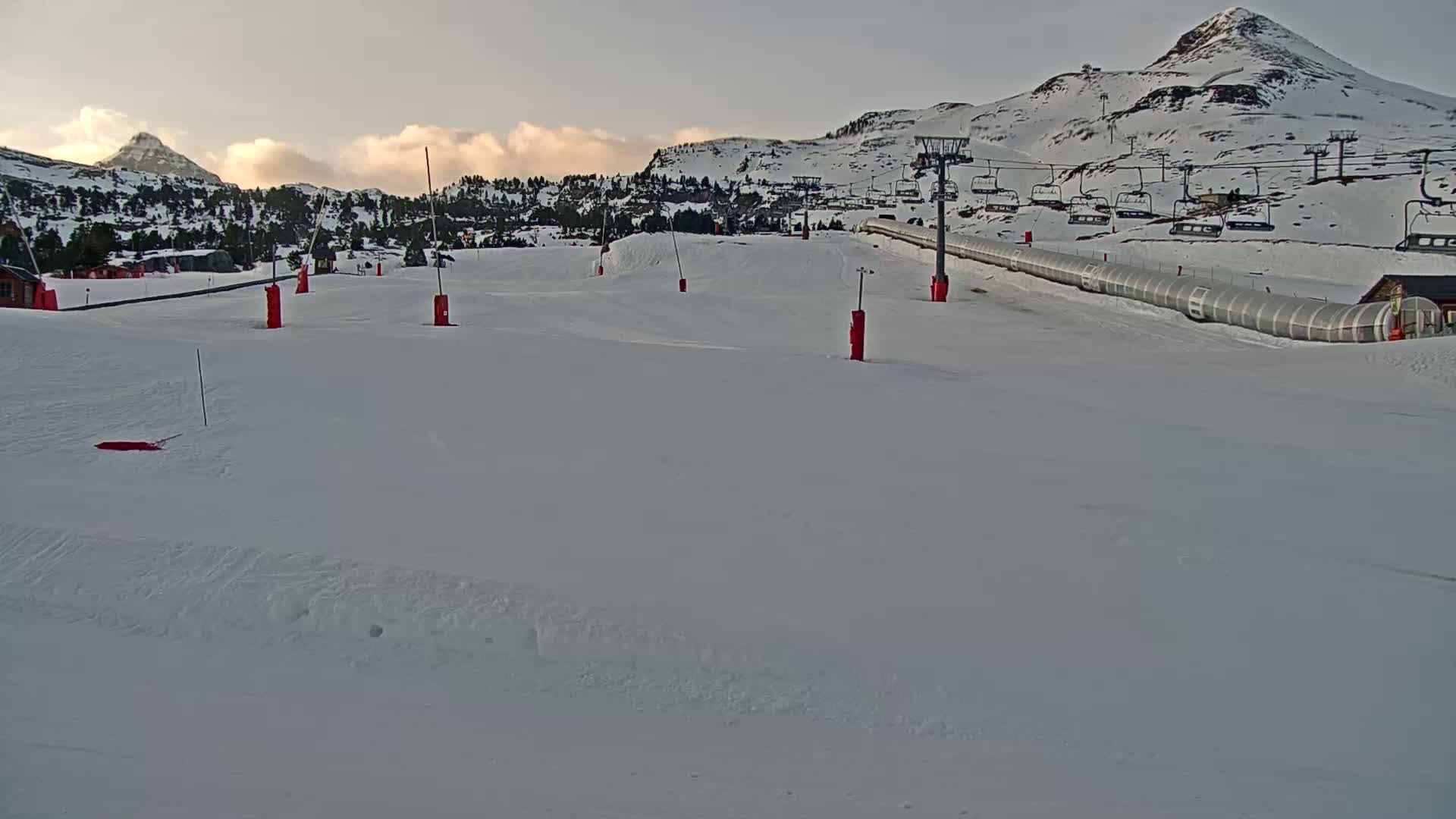 A snow-covered ski slope with a chairlift and a long, covered structure stretches across the lower portion of the scene, all set against a backdrop of snow-dusted mountains under a partly cloudy sky.