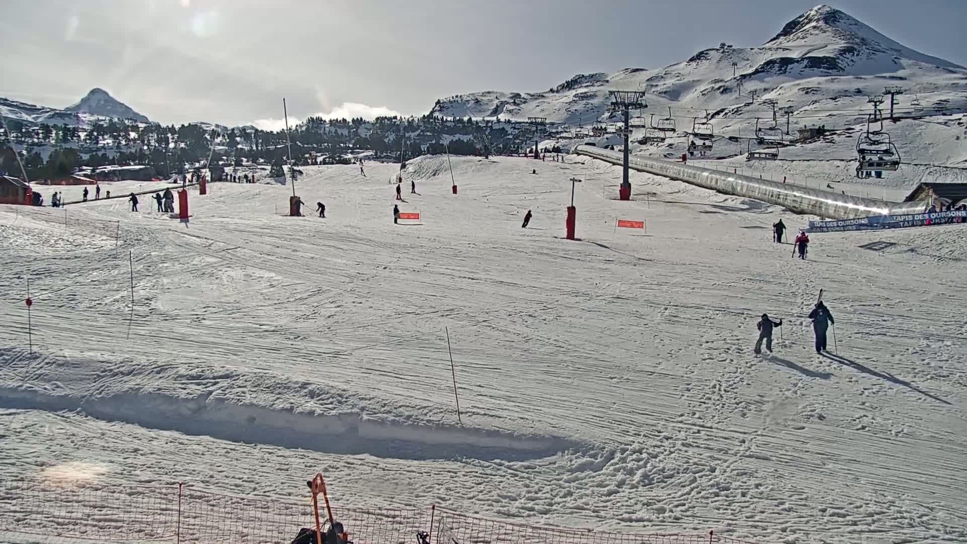 A sunny day at a snow-covered ski resort shows several skiers and snowboarders on the slopes with ski lifts and mountains in the background.