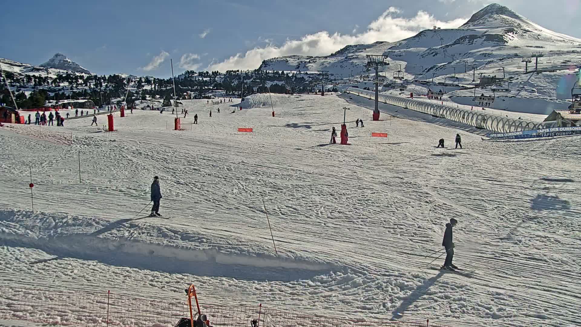 A sunny day on a snow-covered ski slope with several skiers and snowboarders, ski lifts, and snow-covered mountains in the background.