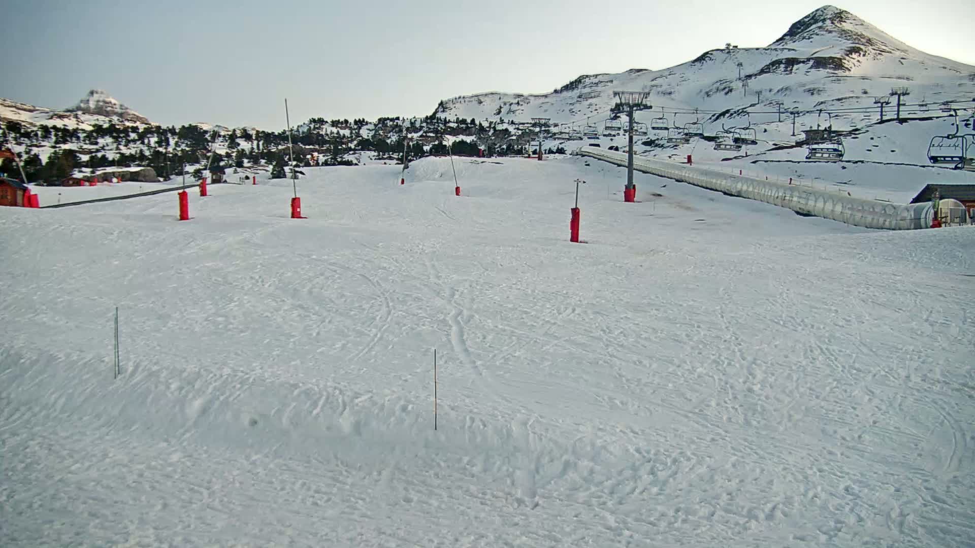 A snow-covered ski slope with several chairlifts and red markers in the foreground, under a clear sky and a snow-capped mountain in the background.