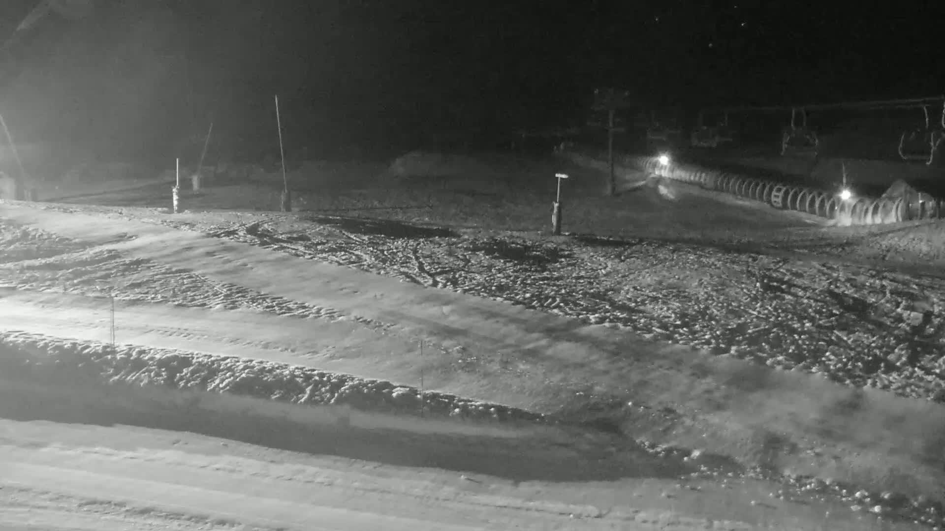 A snow-covered ski slope at night, illuminated by artificial lights, with ski lifts visible in the background.