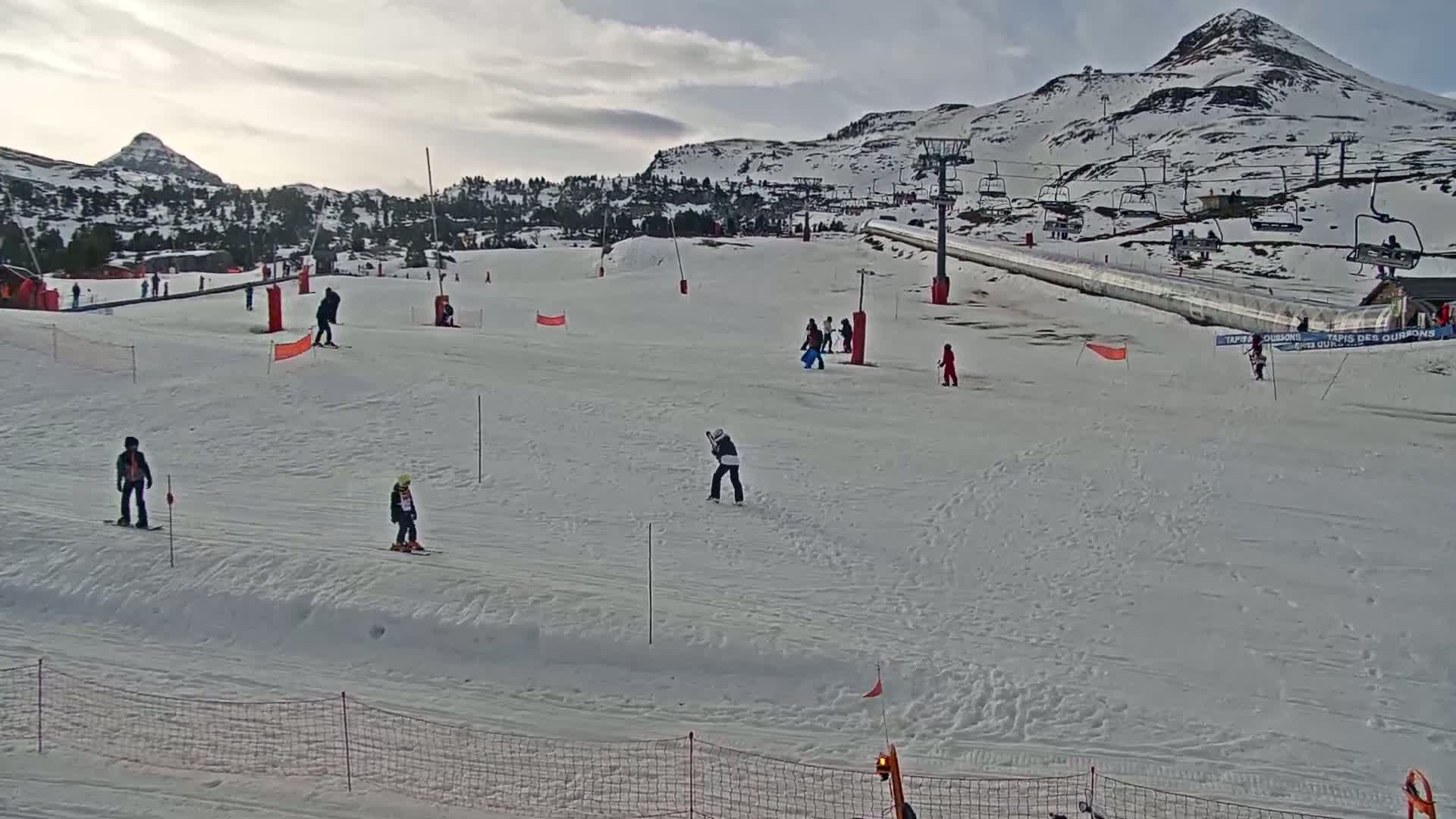 A snow-covered ski slope with several skiers and snowboarders, under a cloudy sky, features a mountain in the background and ski lifts to the right.