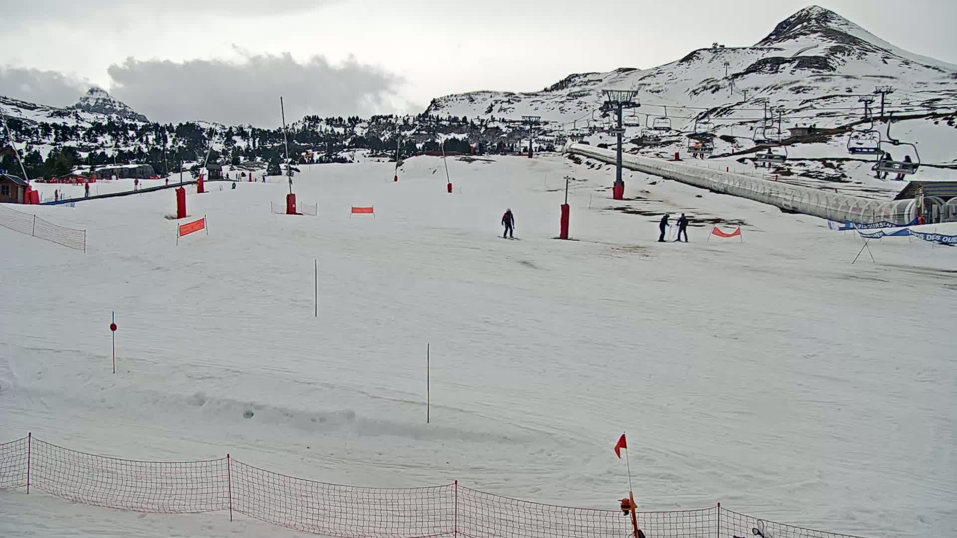 A snow-covered ski slope with several skiers, ski lifts, and snow-covered mountains in the background under an overcast sky.