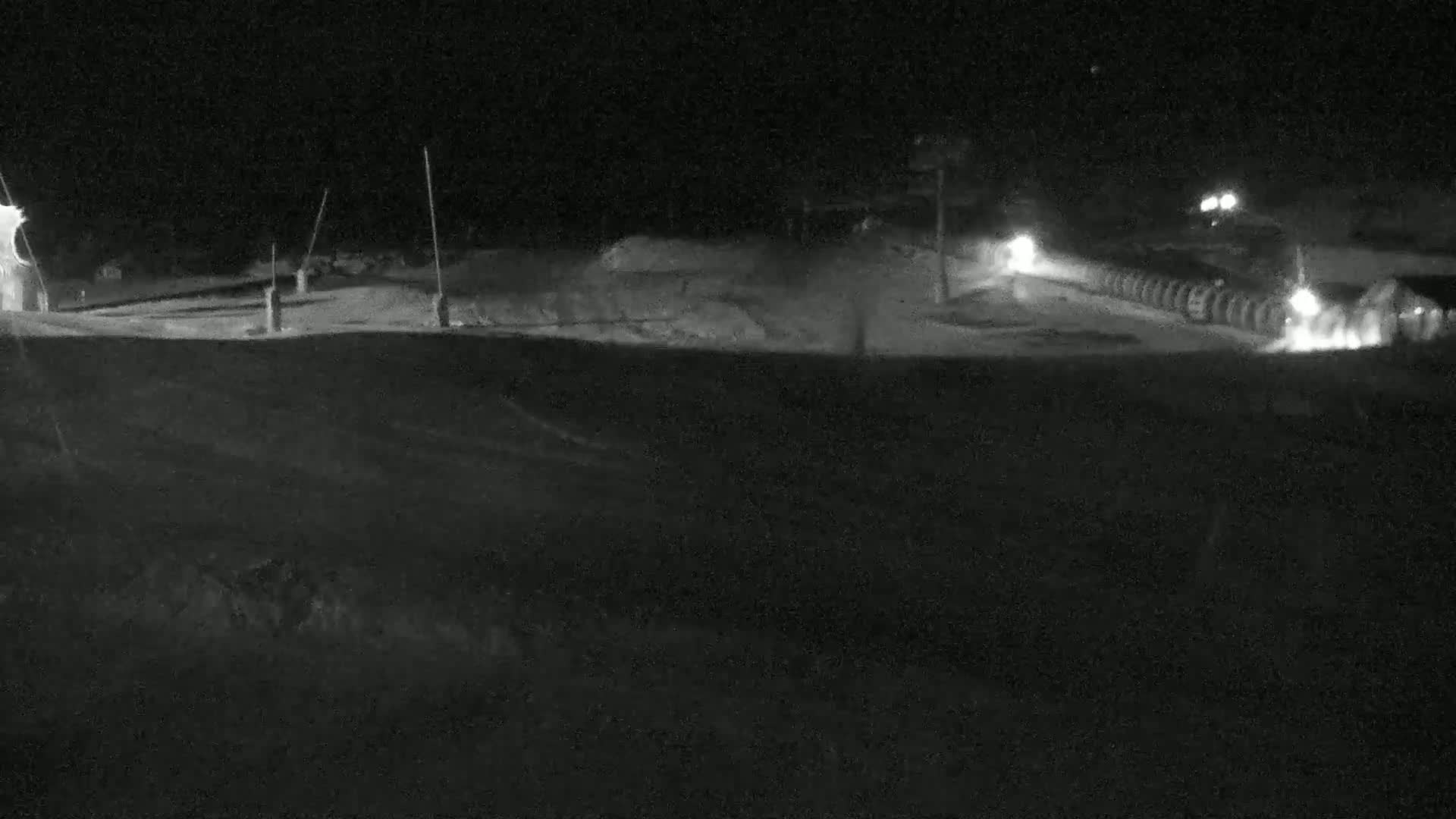 A nighttime, snow-covered scene shows a flat, open area with vehicles and lights in the distance.