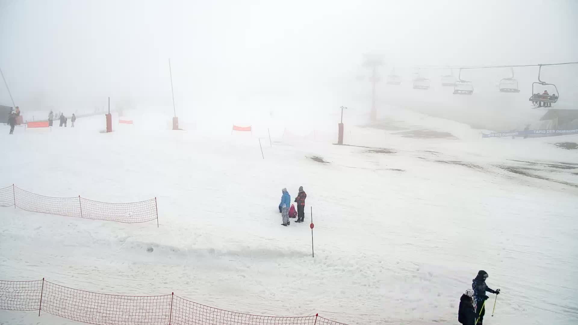 A snow-covered ski slope is shrouded in thick fog, with several people and a chairlift visible in the distance.