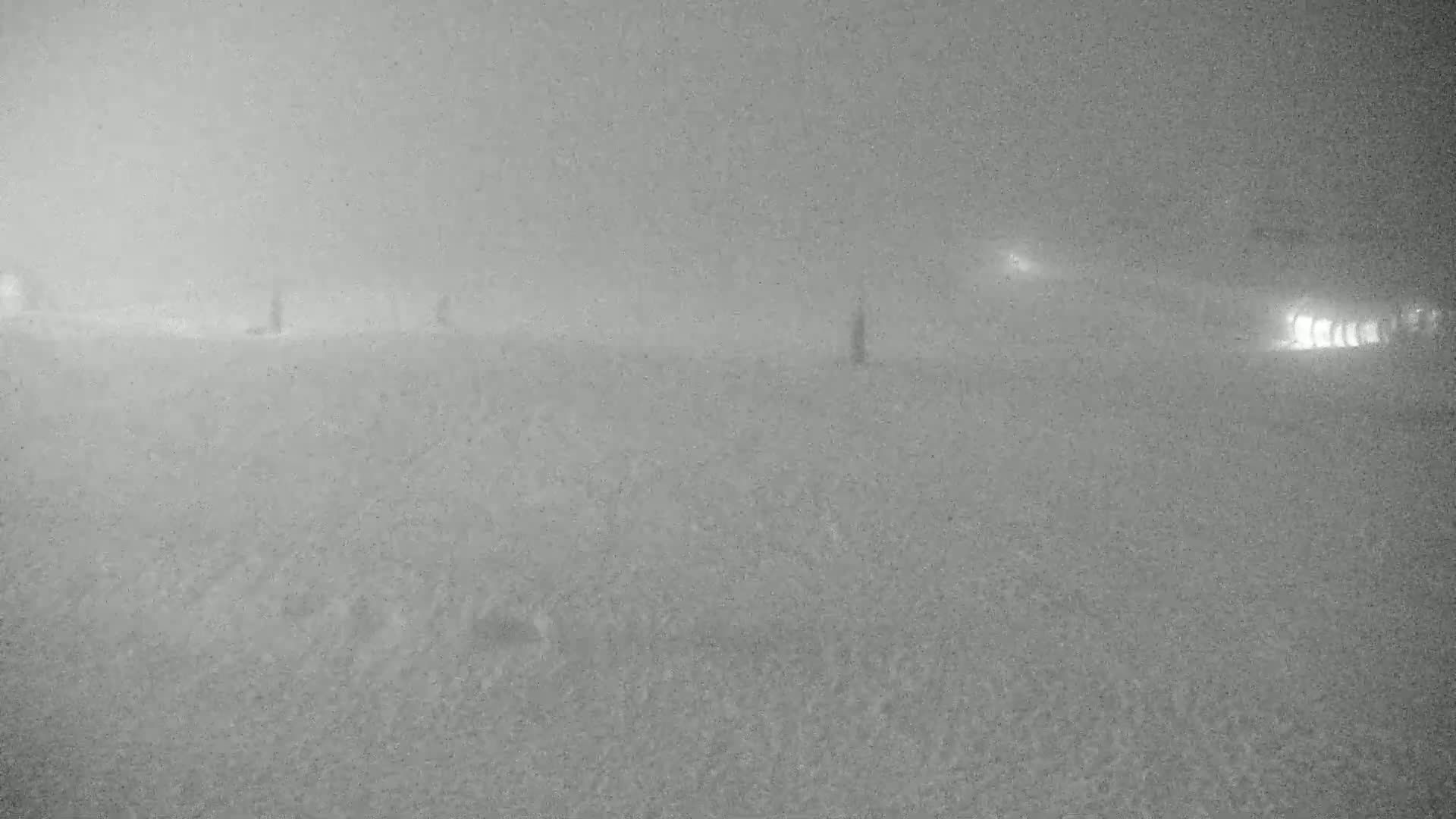 A flat, snow-covered expanse is visible under heavy snowfall with indistinct shapes in the distance and faint lights at the far right.