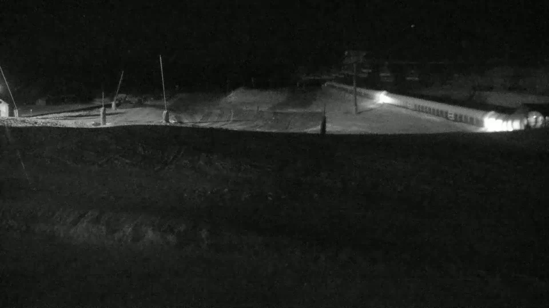 A snow-covered area at night, illuminated by lights from a building, shows what appears to be a ski slope or similar recreational area.