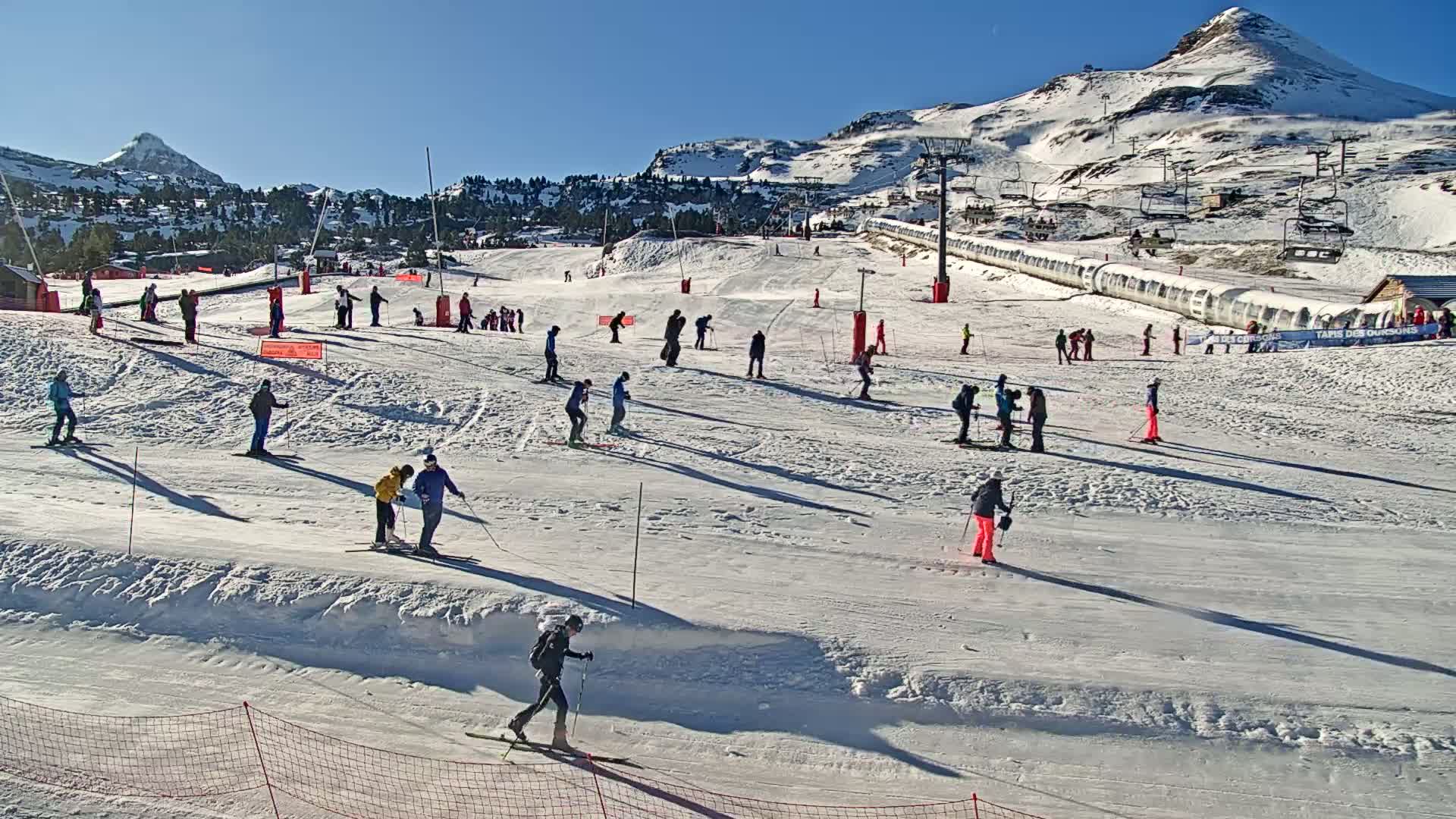 A sunny day on a snow-covered ski slope shows numerous skiers and snowboarders of varying skill levels enjoying the mountain scenery.