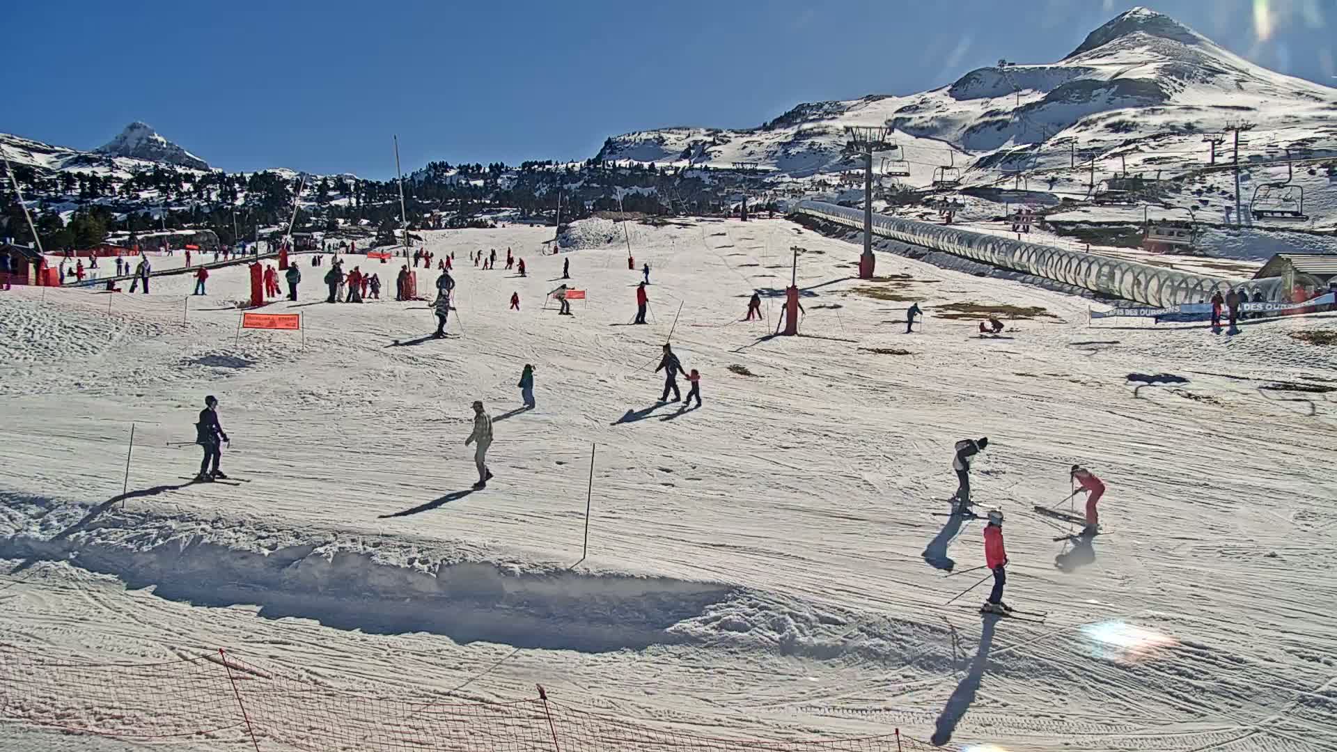 A sunny day at a snow-covered ski resort shows numerous skiers and snowboarders enjoying the slopes, with mountains in the background.