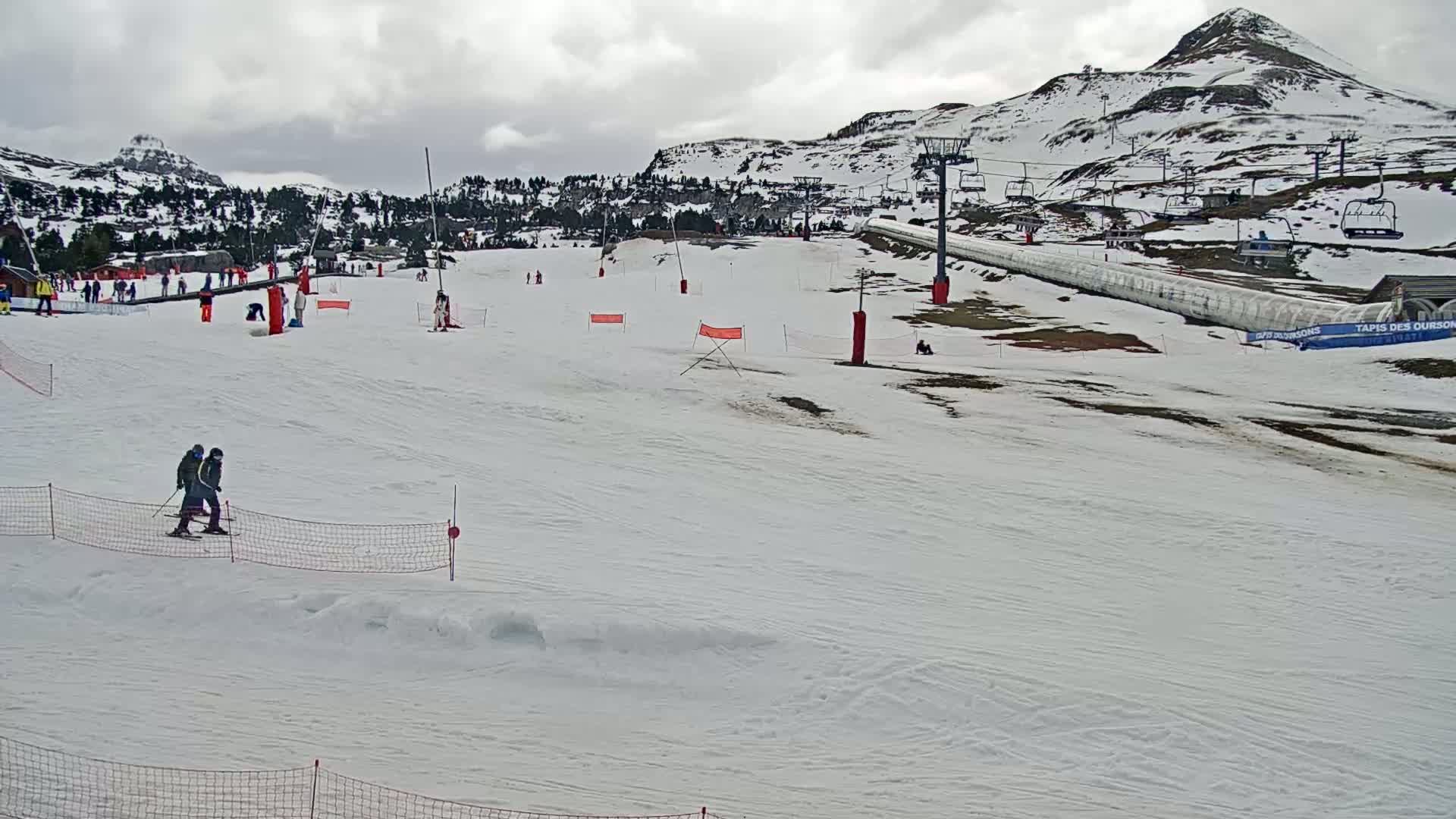 A snowy ski slope with several skiers and snowboarders, ski lifts, and a mountain in the background under an overcast sky.